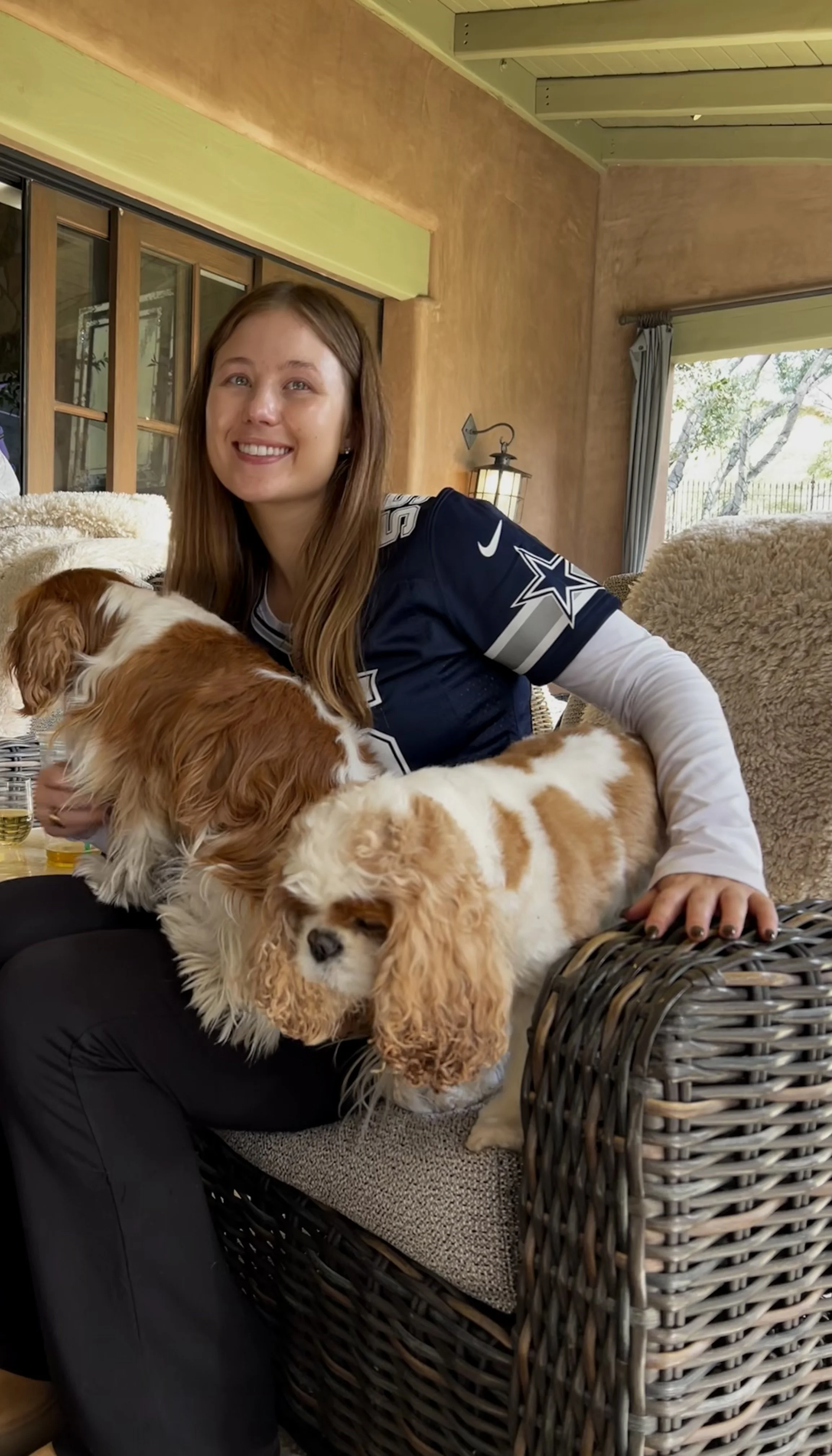 A woman with long brown hair, wearing a Dallas Cowboys football jersey, sitting on a wicker chair with two adorable Cavalier King Charles Spaniel puppies, one lying on her lap and the other close on the chair.