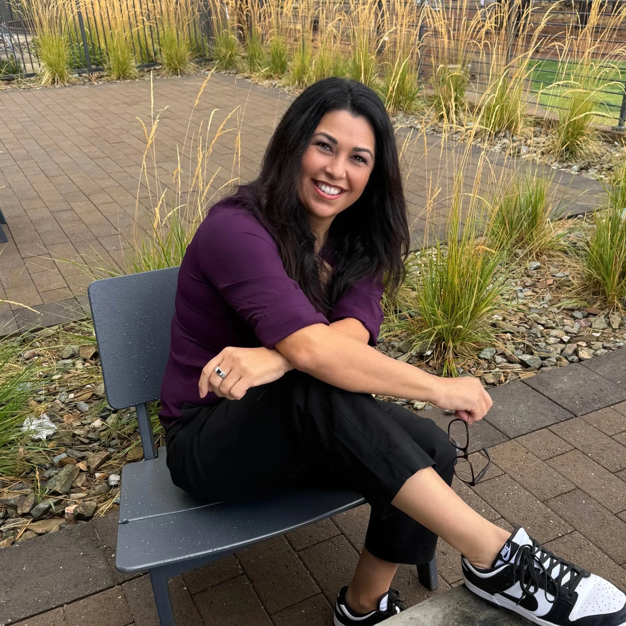 A woman with long dark hair, smiling, sitting on a bench outdoors with plants and a brick walking path in the background. She is wearing a purple top and black shorts, and has sneakers on.