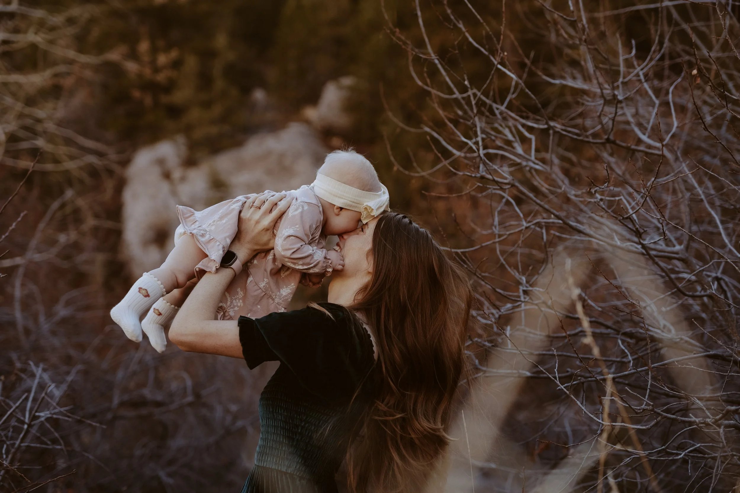 Woman lifting child in a wooded outdoor area during fall, both sharing a kiss.