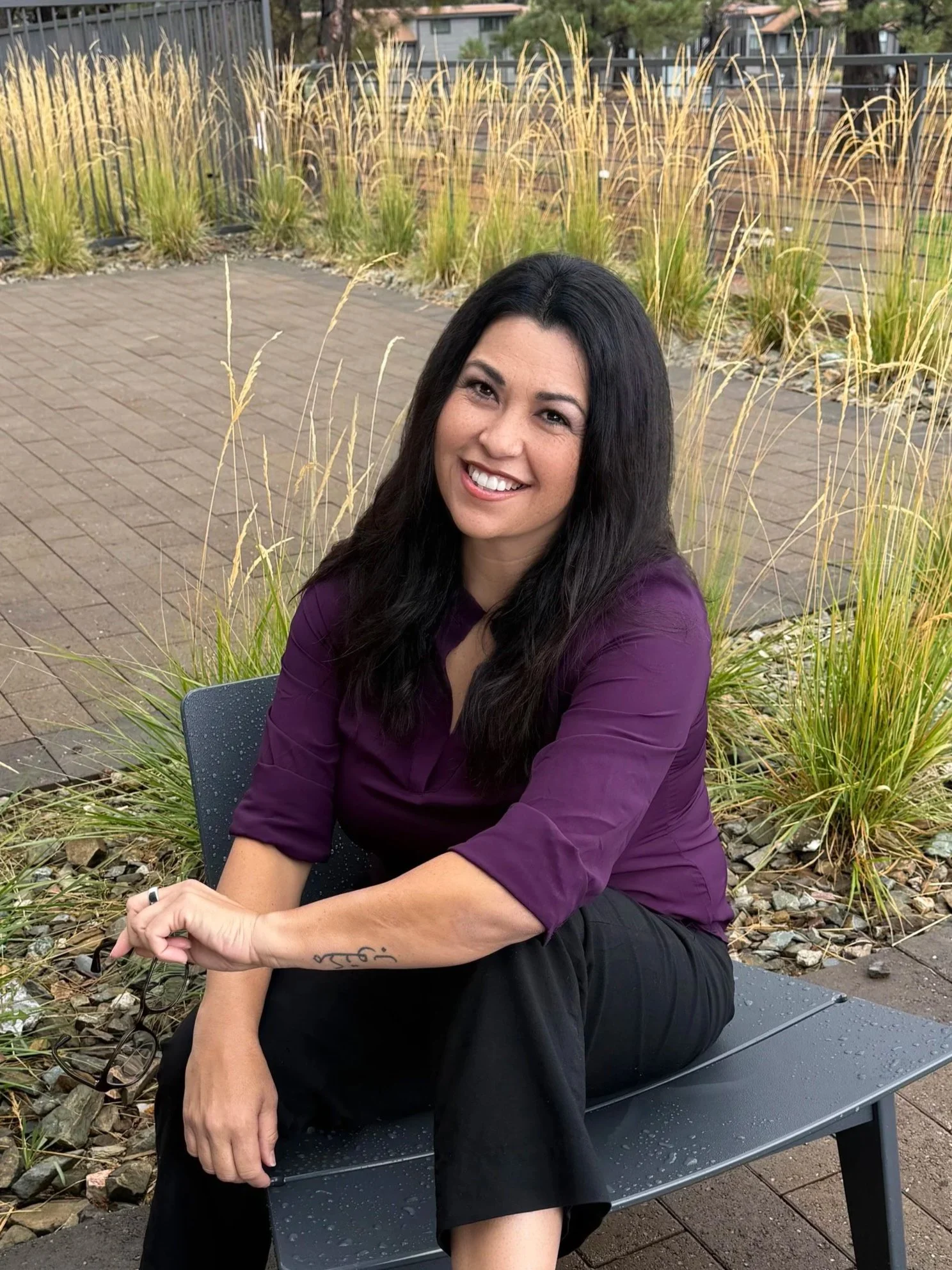A mental health therapist woman with long dark hair, smiling, sitting on a black bench outdoors in front of yellow ornamental grasses and buildings in the background, wearing a purple top and black pants.