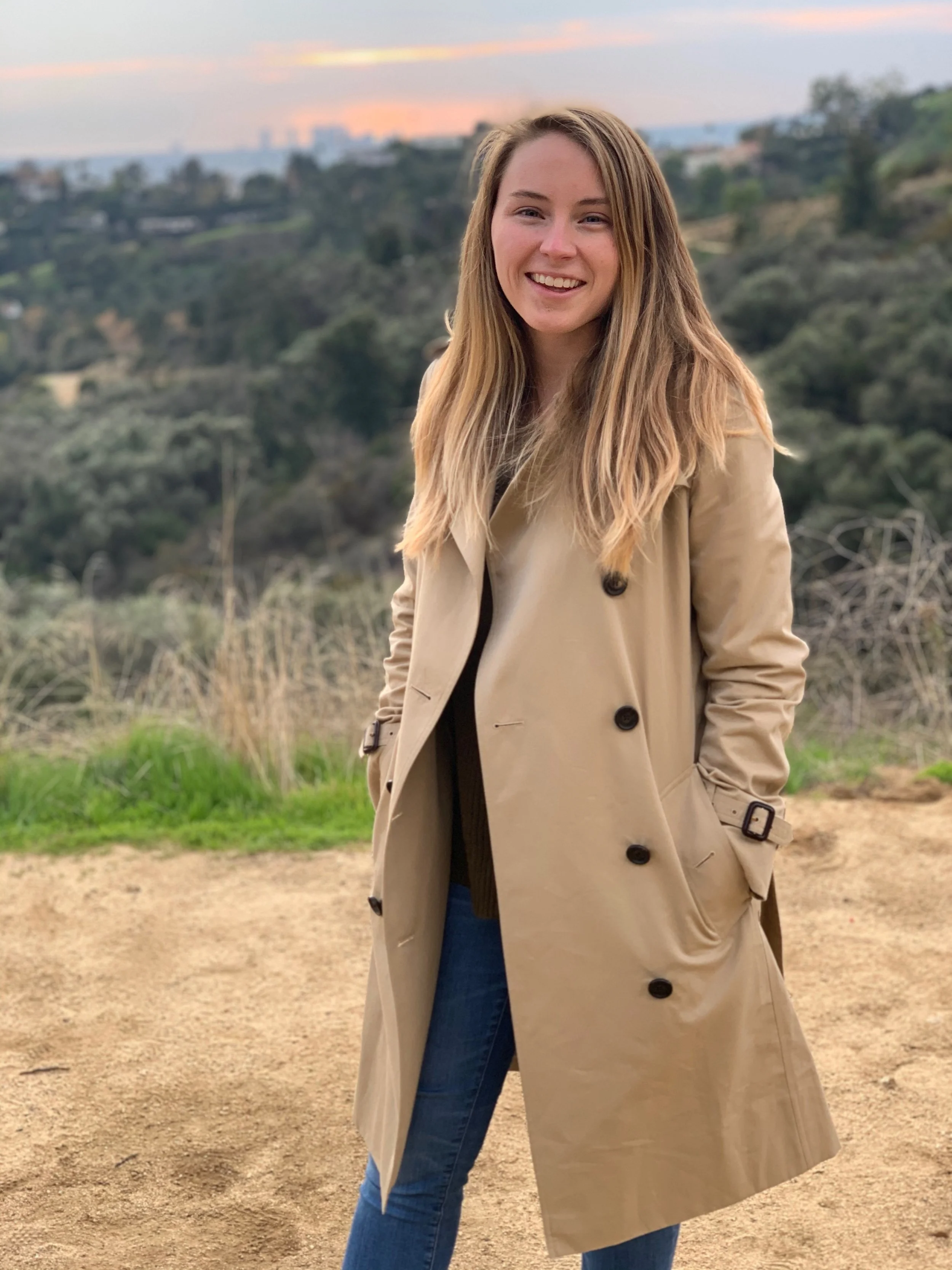 A young woman standing outdoors on a dirt path, smiling, wearing a beige trench coat and blue jeans, with a scenic hilly landscape and a sunset sky in the background.