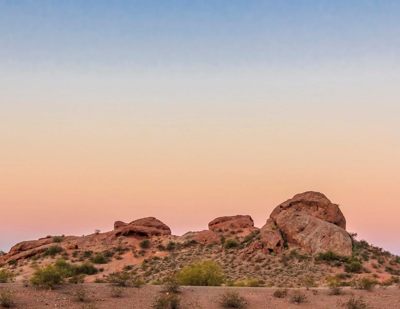 Papago Peak, a rocky outcropping in red and orange tones, rises above a desert landscape. At the top of the image, a dusky sky of blue, yellow, orange, and pink loom over the peak.
