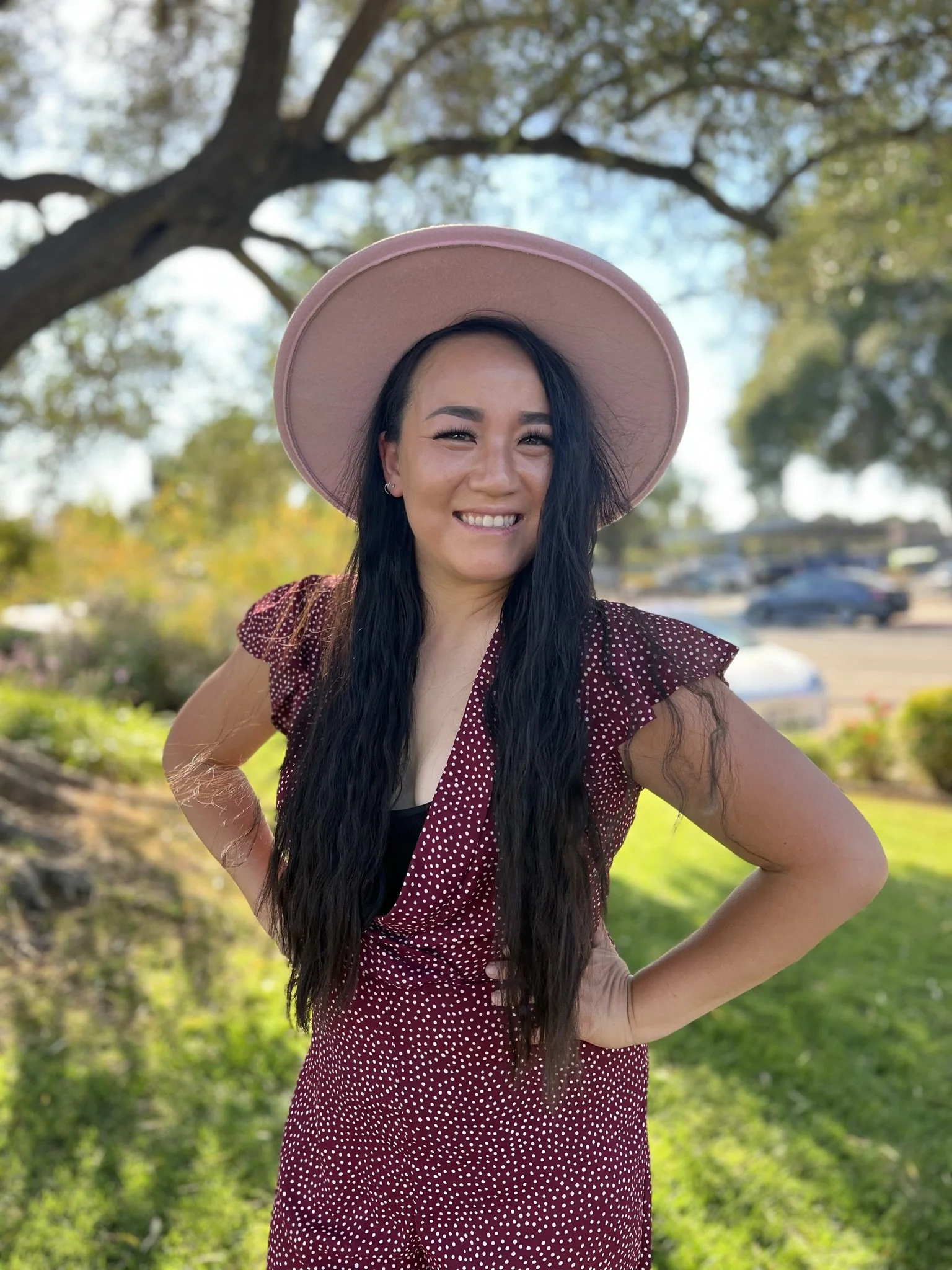 A woman smiling outdoors, wearing a wide-brimmed pink hat and a maroon dress with white polka dots, standing in front of a tree with green foliage and a parking lot in the background.