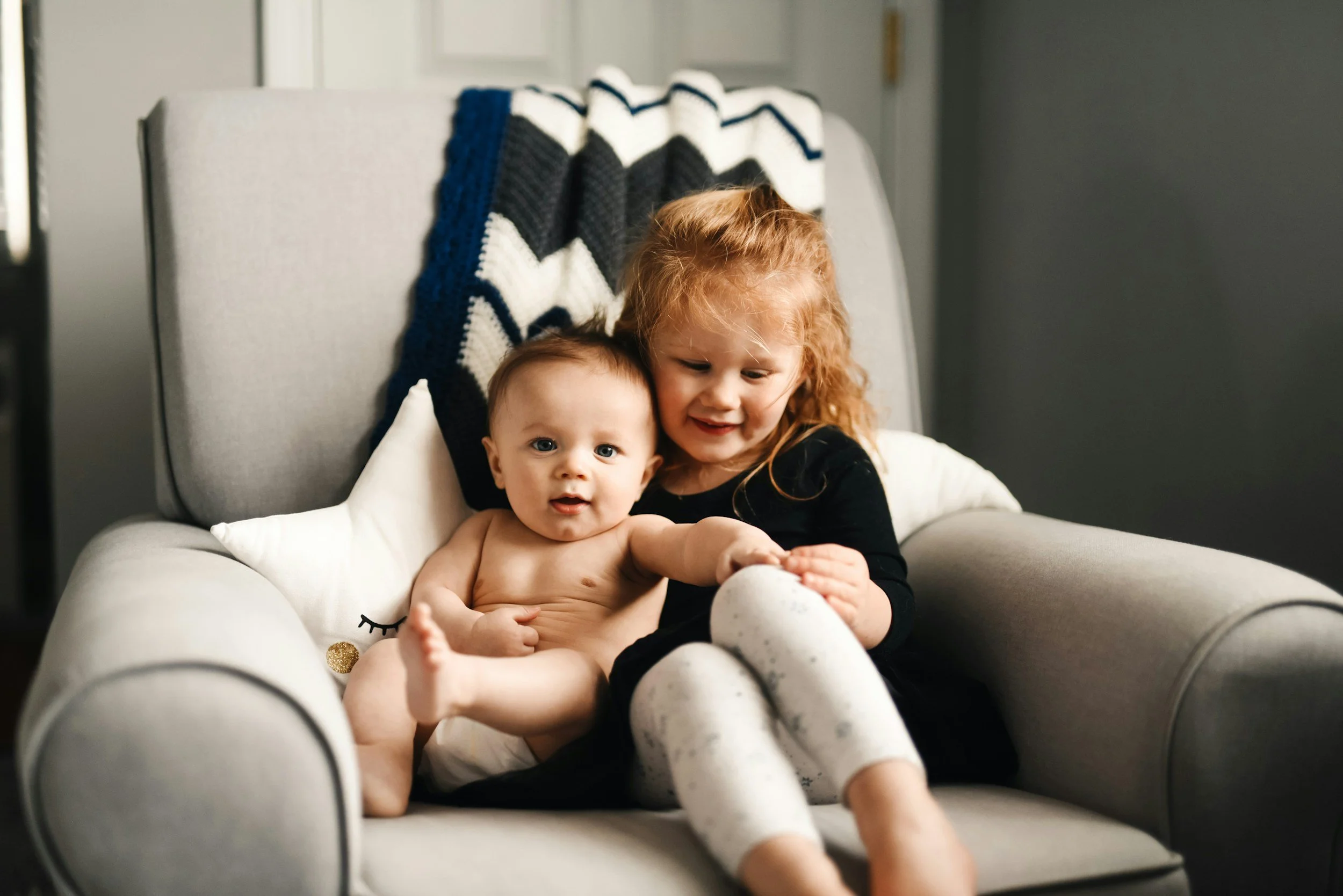 A young girl with red hair and pajamas sitting on a gray armchair, holding a baby boy with light skin and no clothes. The girl is smiling and looking at the baby, who is looking at the camera.