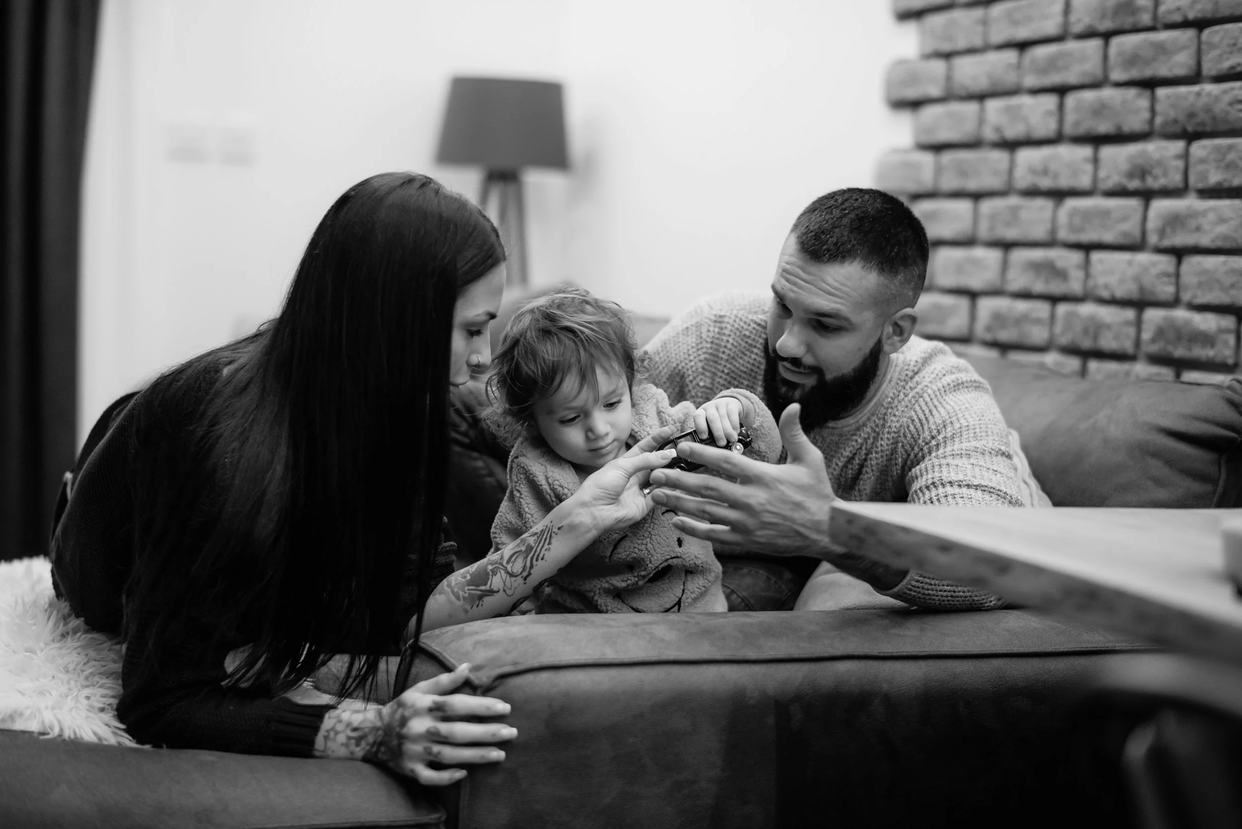 A family of three, including a woman with tattoos, a man, and a young child, are sitting on a couch looking at a phone together in a cozy living room with a brick wall and a lamp in the background.