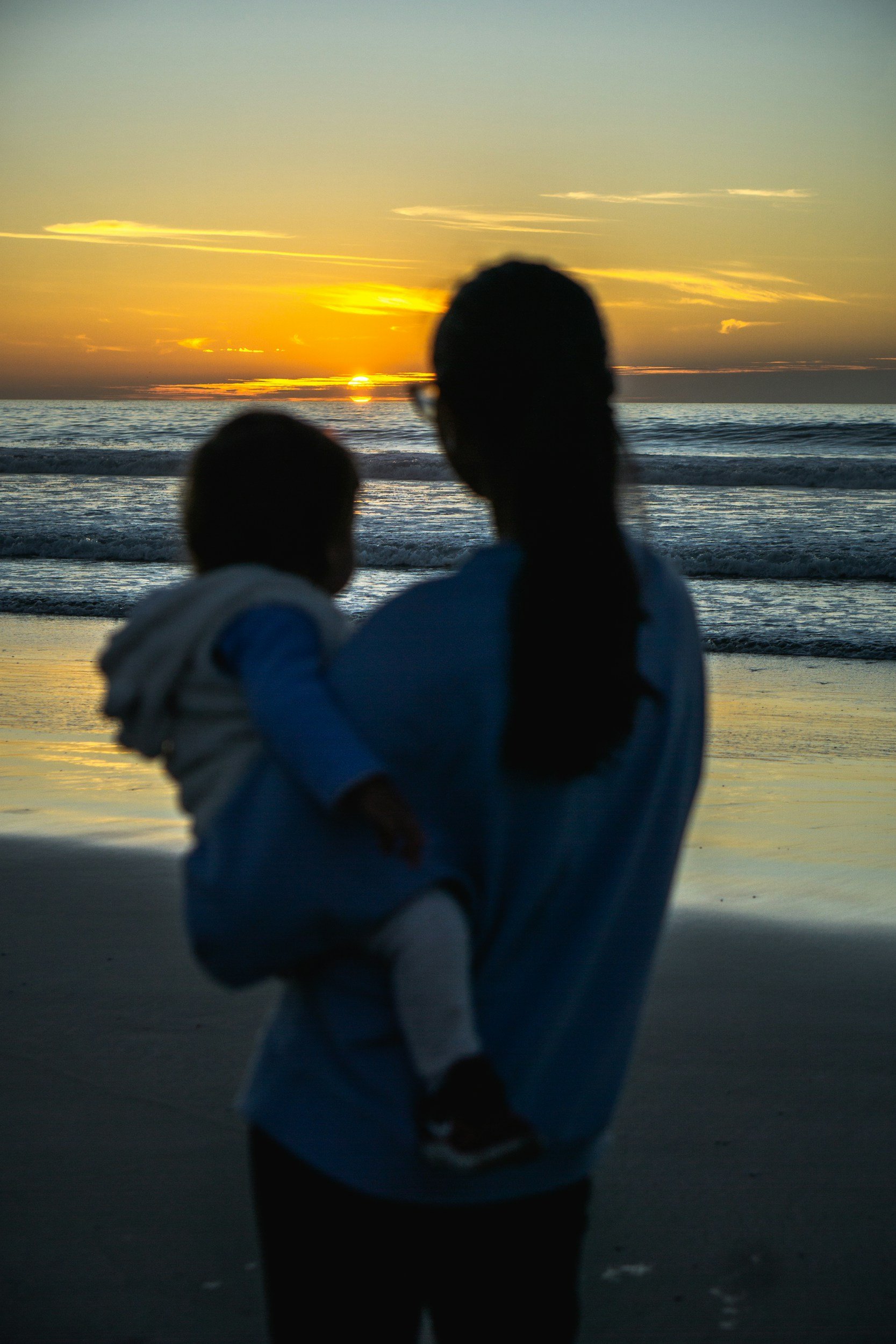 Silhouette of a woman holding a child on her hip, looking at a sunset over the ocean at the beach.