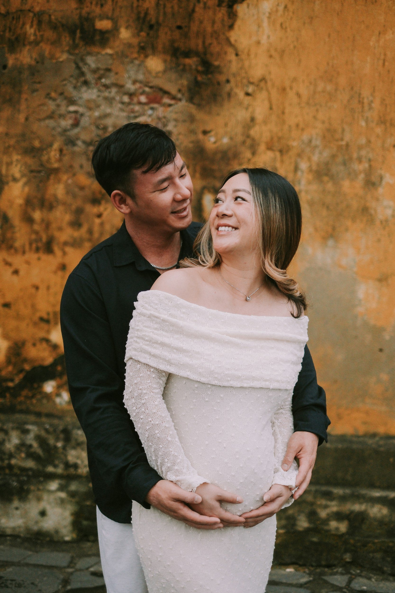A happy pregnant woman in a white dress with off-shoulder sleeves stands in front of a textured orange wall, smiling at a man behind her who is holding her belly. They are smiling, looking into each other's eyes.