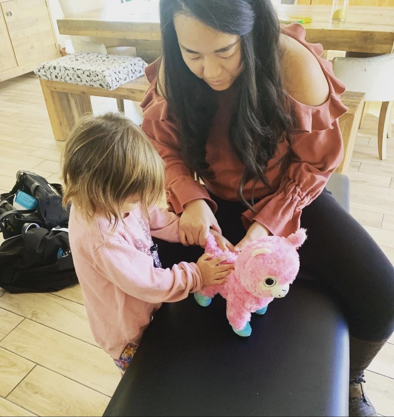 A woman and a young girl are playing with a pink plush llama toy in a cozy room with wooden furniture and a wooden floor.