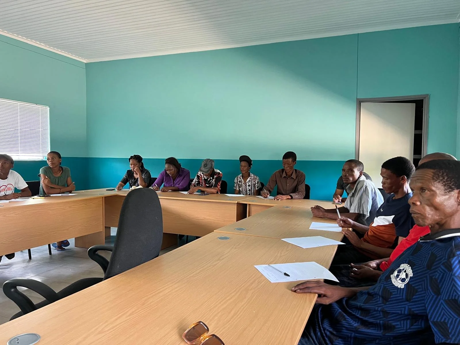 People sitting around a U-shaped conference table in a room with teal walls, some taking notes, listening, and engaging in a meeting or discussion.