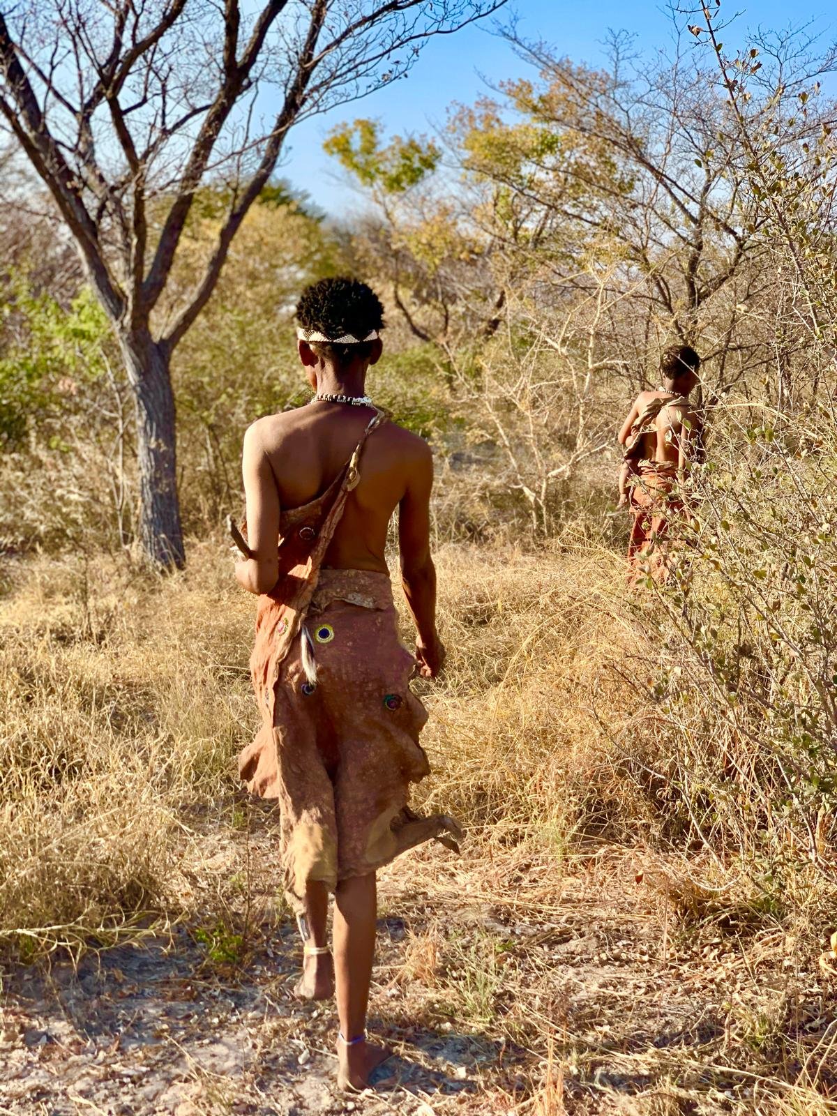 Three individuals walk through a dry, grassy landscape with scattered trees and bushes. They are dressed in traditional or tribal attire, with minimal clothing and decorative accessories, and their hair is styled distinctive to their culture.
