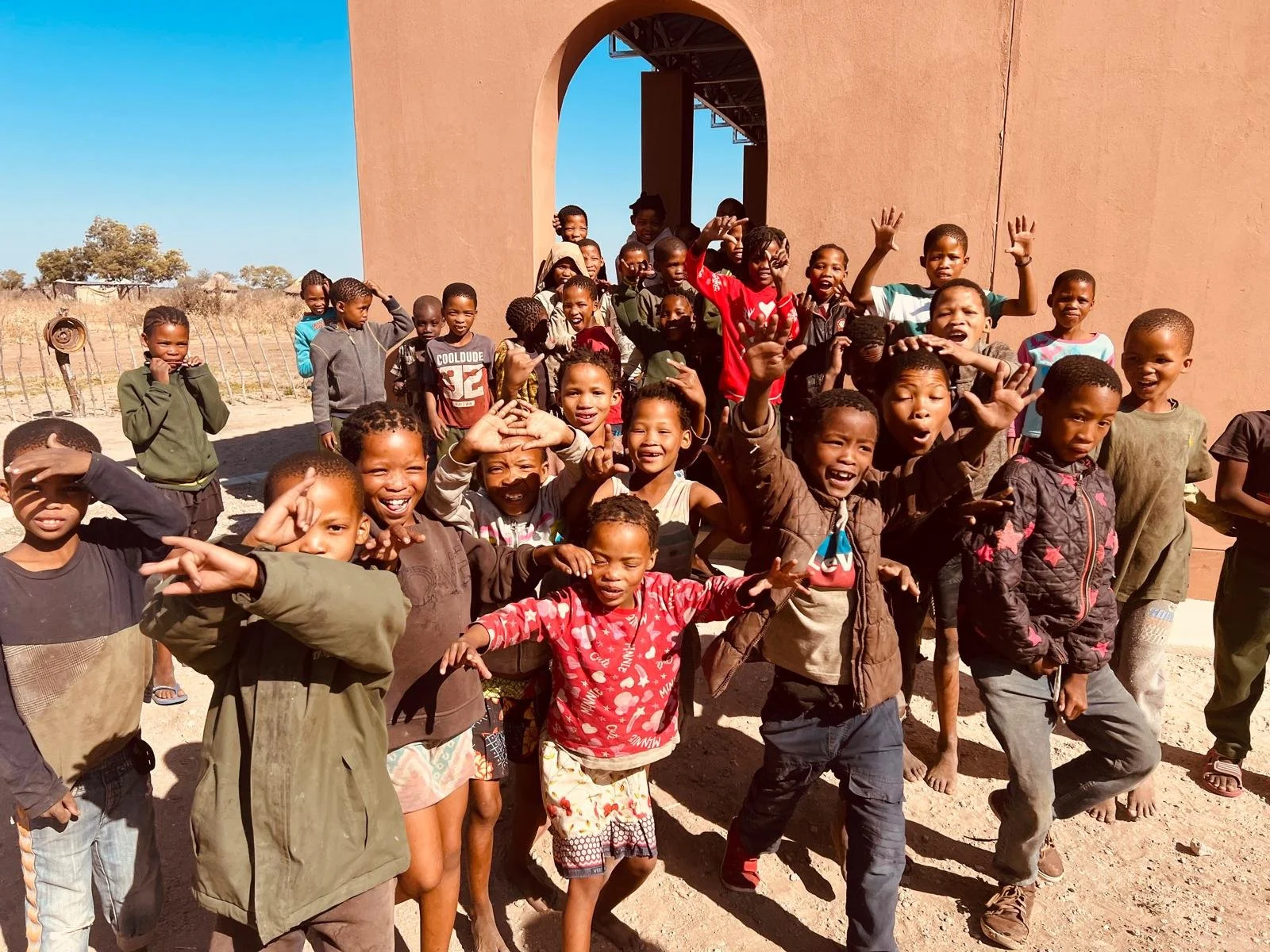 A group of smiling children and a few adults standing outside near a building with a large arched opening in a rural setting on a sunny day.