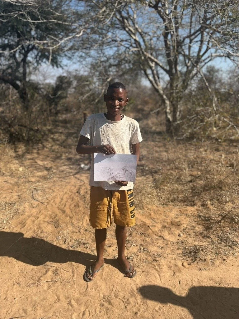 A young boy standing outdoors on a dirt path, holding a drawing of trees and a landscape, with leafless trees and a clear blue sky in the background.