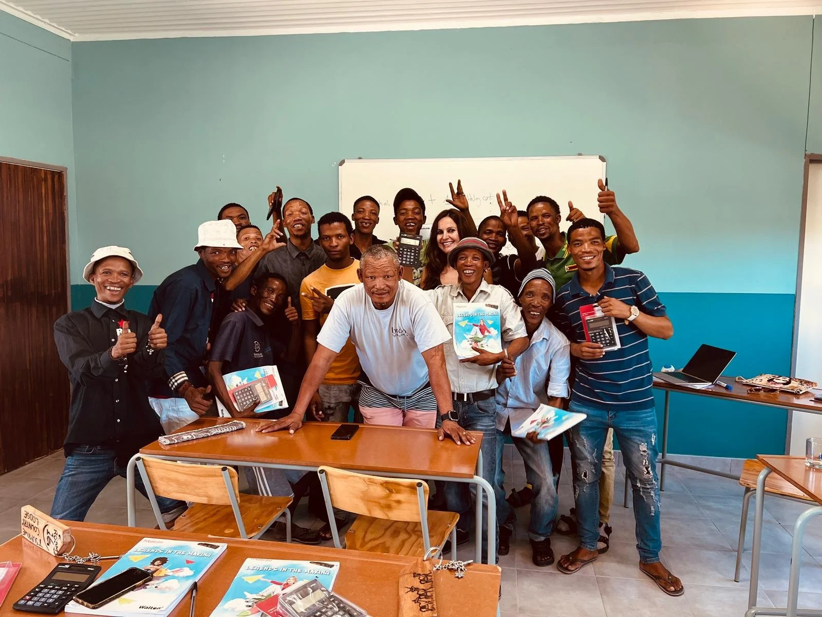 Group of smiling adults and children in a classroom, holding books and calculators, posing for a photo.