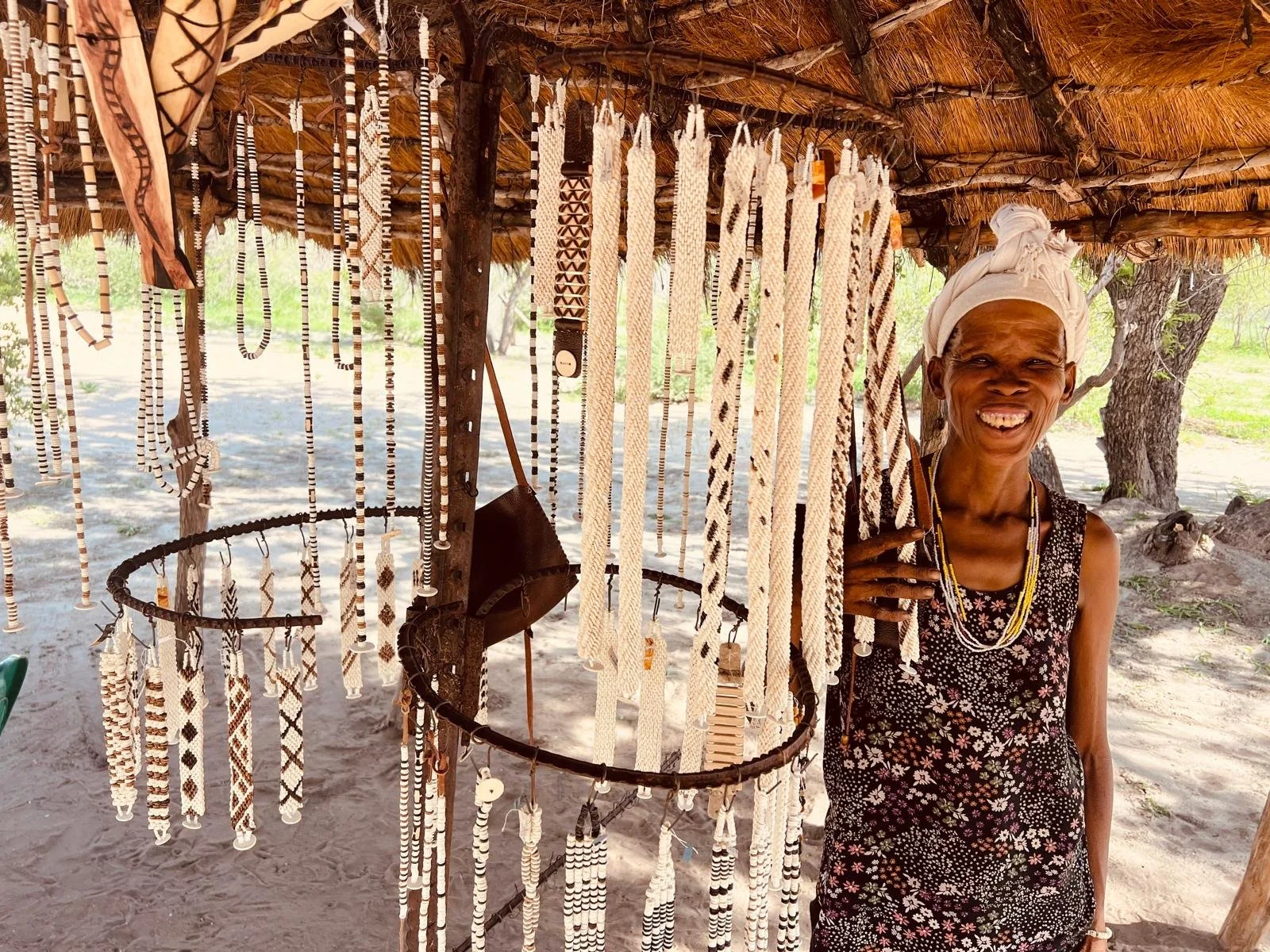 A smiling woman standing behind a display of handmade beaded jewelry at a market stall in an outdoor setting with trees in the background.
