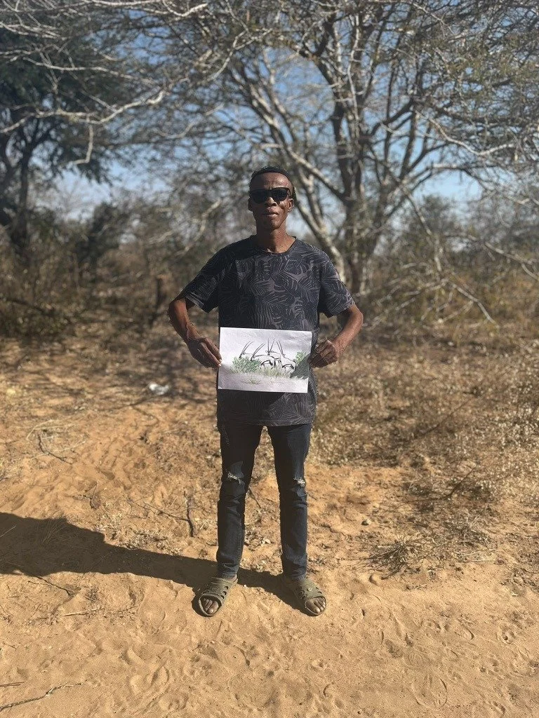 Man standing outdoors on dirt ground holding a drawing of grass and plants, wearing sunglasses, dark shirt, ripped jeans, and sandals, with leafless trees in the background.