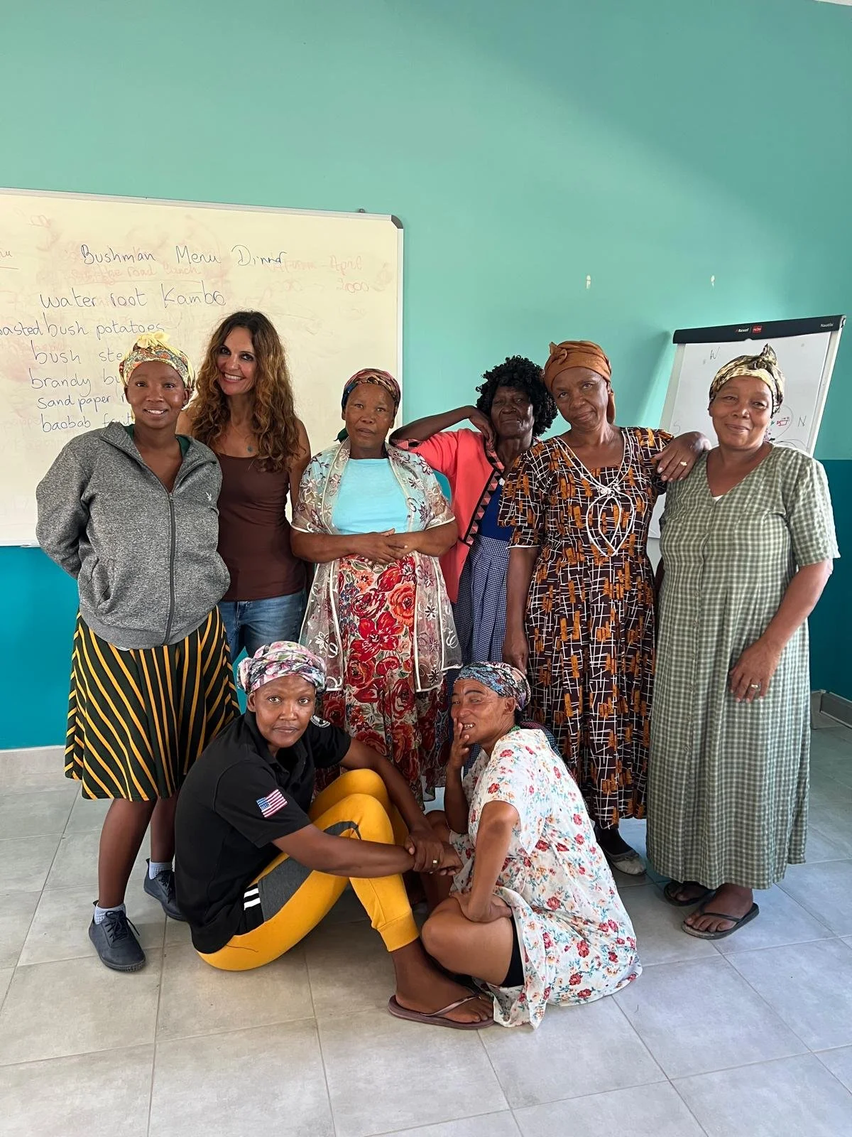 A group of nine women and one man posing together in a classroom with a whiteboard and a teal wall in the background. Some women are standing, one woman and a man are sitting on the floor, and one woman is kneeling. They are wearing various colorful and patterned clothing, and some women are wearing headscarves.