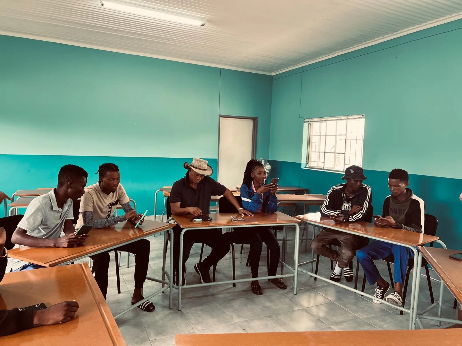 Group of six young people sitting at desks in a classroom, all looking at their smartphones.