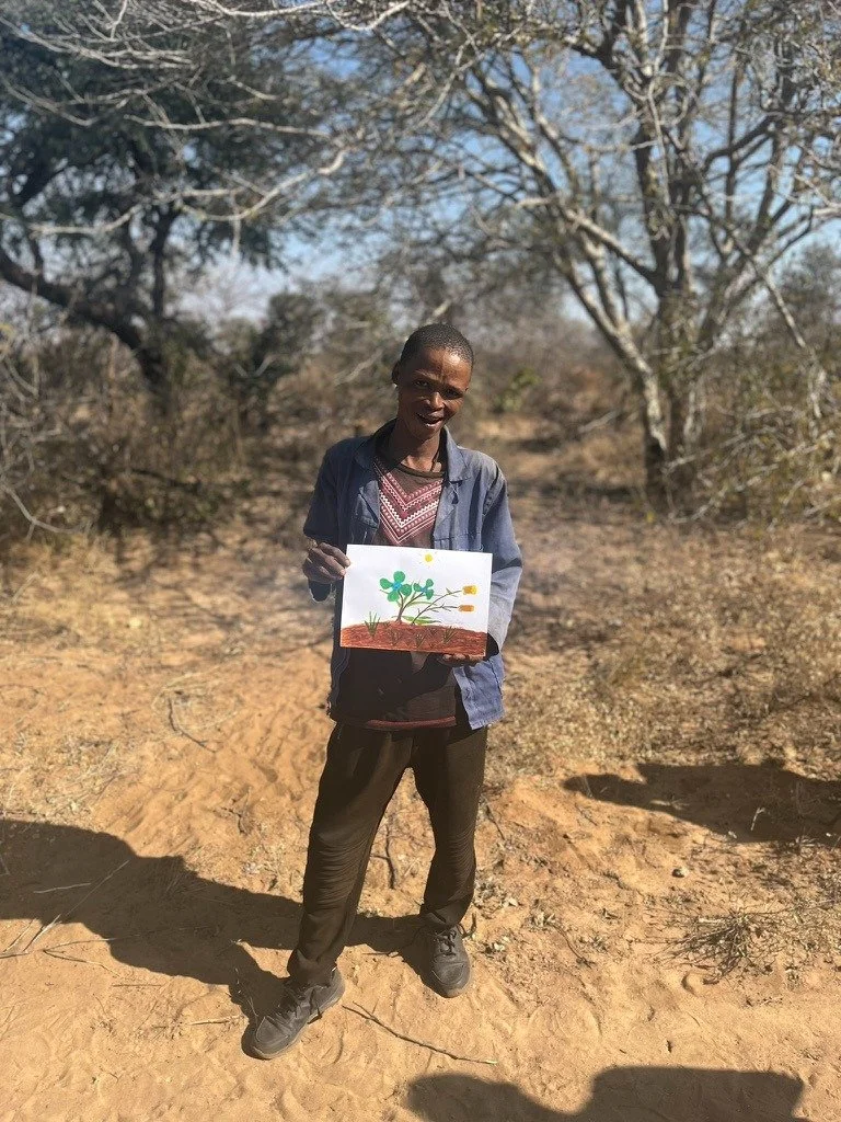 A young person standing outdoors in a dry, bare landscape with leafless trees in the background, holding a drawing of a plant with green leaves and roots.