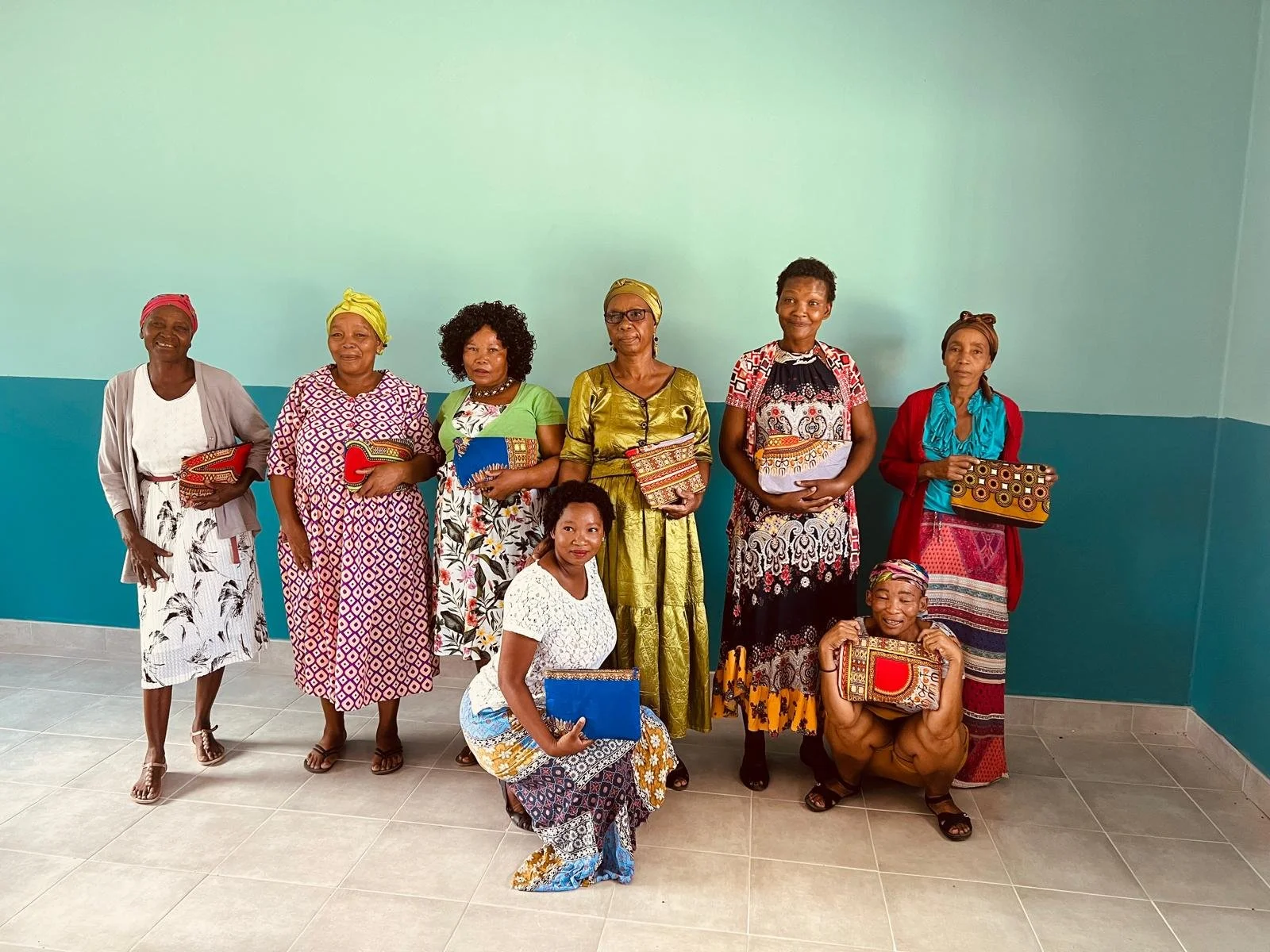 Group of women in colorful dresses holding patterned handbags and folders, posing indoors against a light blue wall.