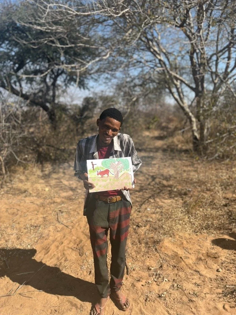 Young man standing outdoors on dirt ground, holding a colorful drawing of a lion, tree, and grass under a blue sky, with leafless trees and bushes in the background.