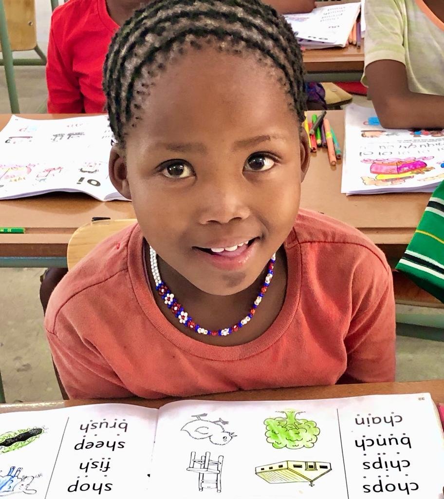 A young girl with braided hair, wearing a red shirt and a beaded necklace, smiling at the camera. She is seated at a desk with an open educational book in front of her in a classroom setting.