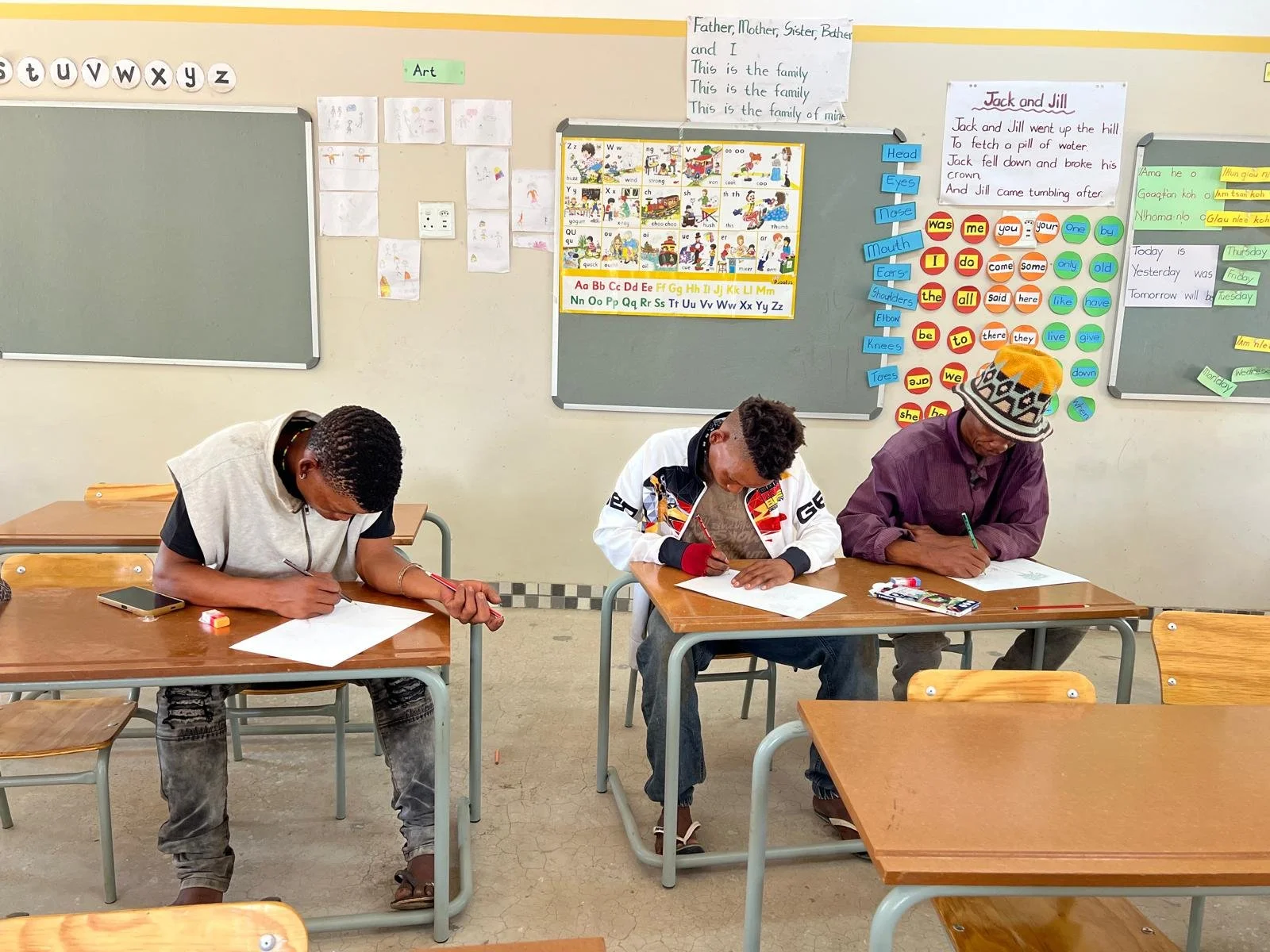Three students sitting at desks in a classroom, writing on paper. The classroom has educational posters and charts on the walls.