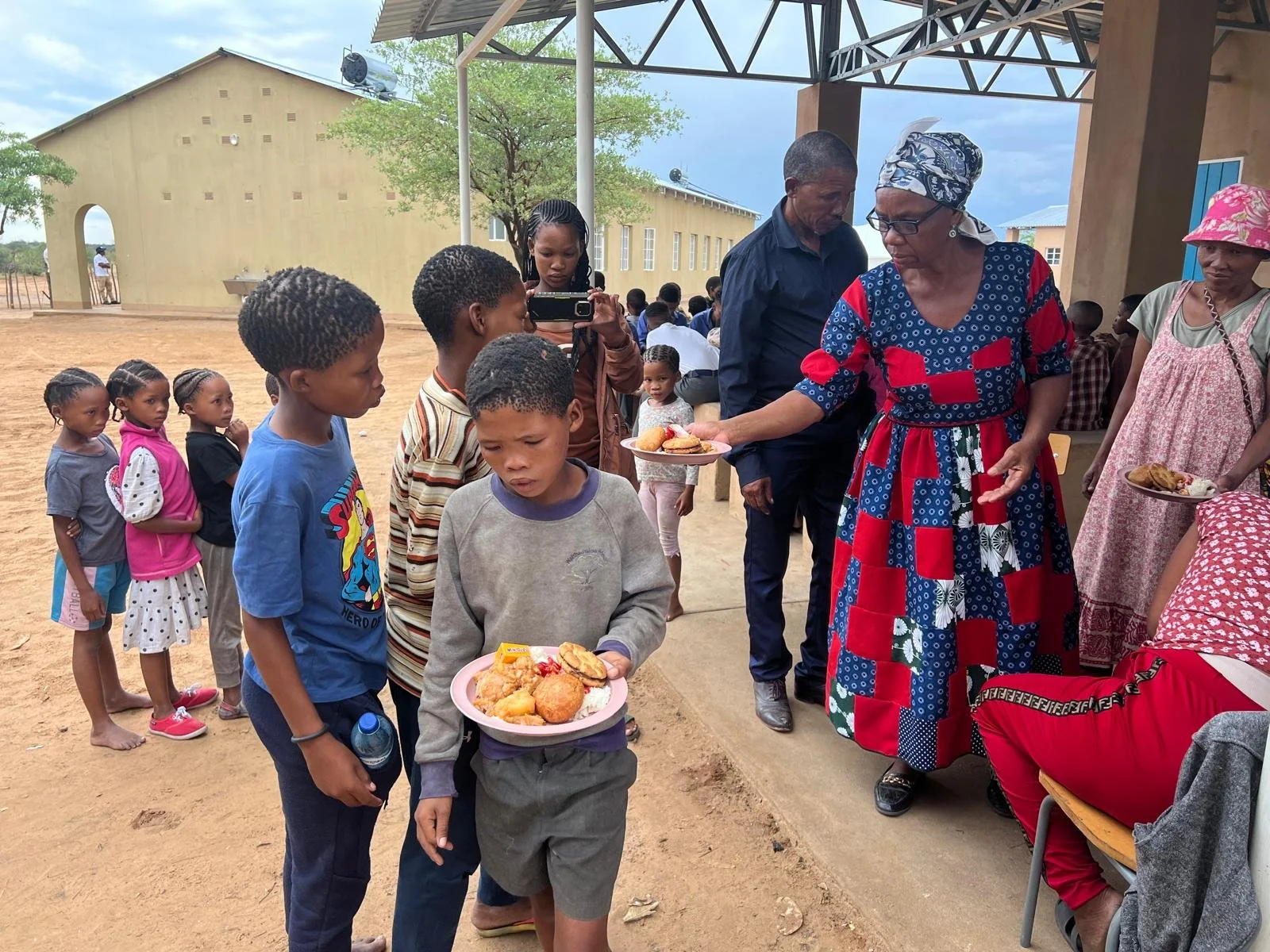 Children in line receiving plates of food at a community gathering outside a building in a rural area.
