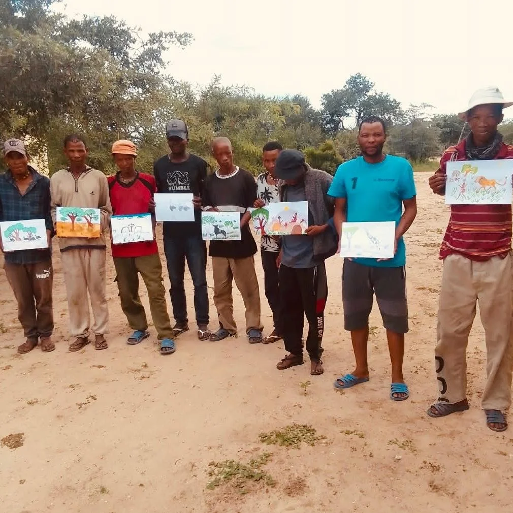 A group of nine people standing outdoors in a line, each holding a colorful drawing or painting of animals and nature scenes.