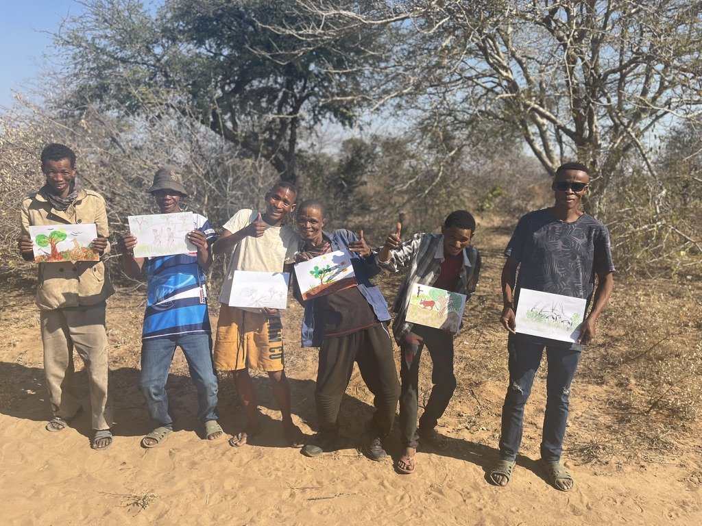 Group of six young men standing outdoors on a dirt path, holding up drawings of trees, animals, and nature scenes with dry trees and clear sky in the background.
