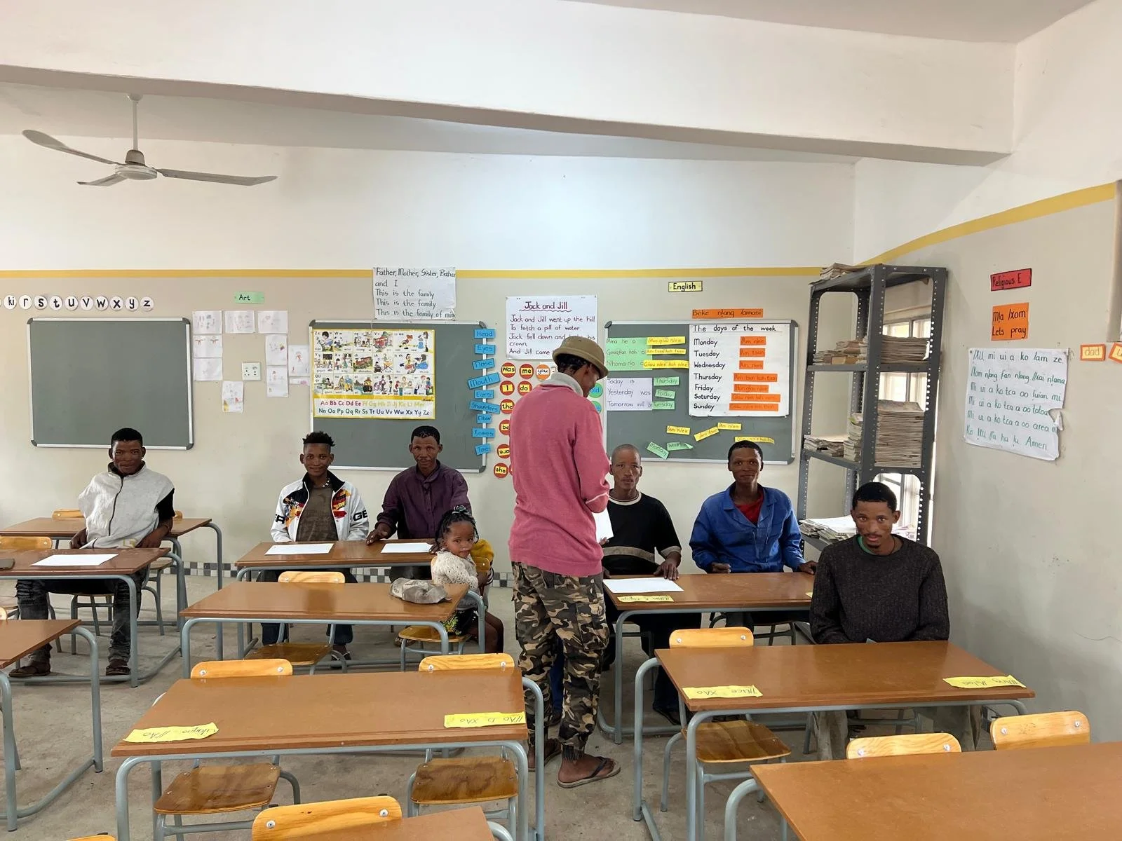 A classroom with seven students seated at desks facing the camera and a man in a pink shirt and cap standing in the middle, looking down. The classroom has educational posters on the walls, a metal shelf with books, and desks with yellow labels.