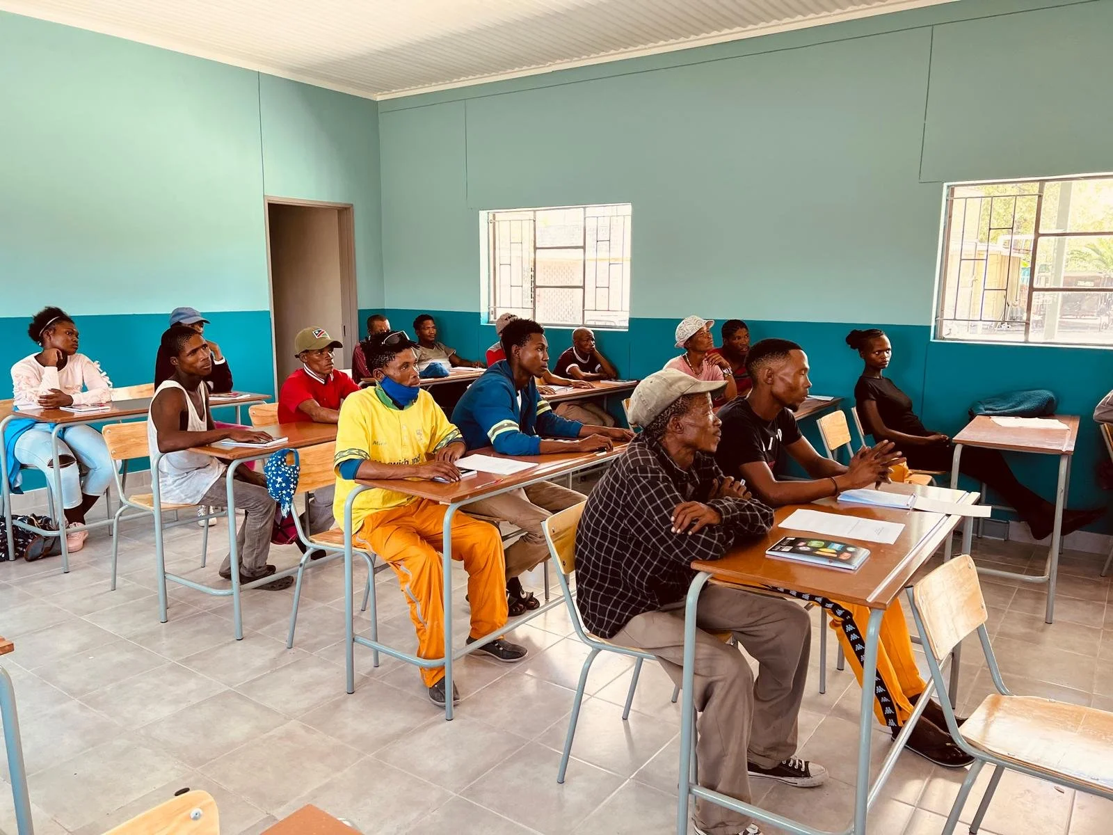 A classroom with students sitting at desks, listening attentively. The room has light green walls, large windows, and tiled floors.