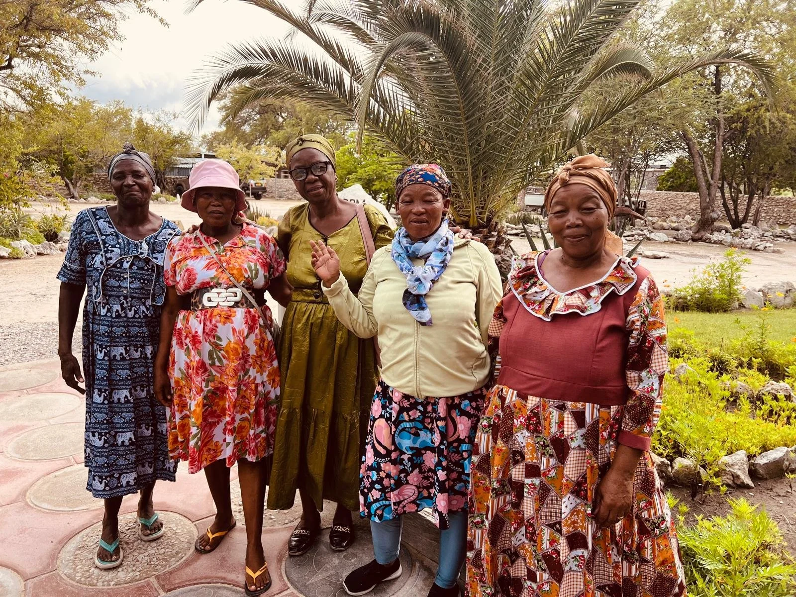 Five women standing outdoors in a garden, posing for a photo, with trees and landscaping in the background.