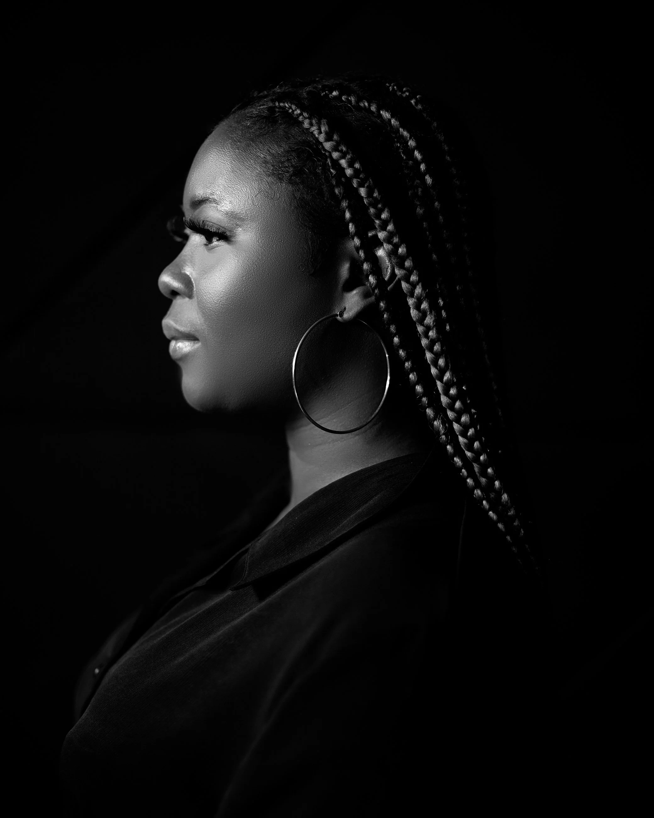 Febe's Profile picture; an African woman with braided hair, wearing large hoop earrings, looking to the left, in black and white with a dark background.
