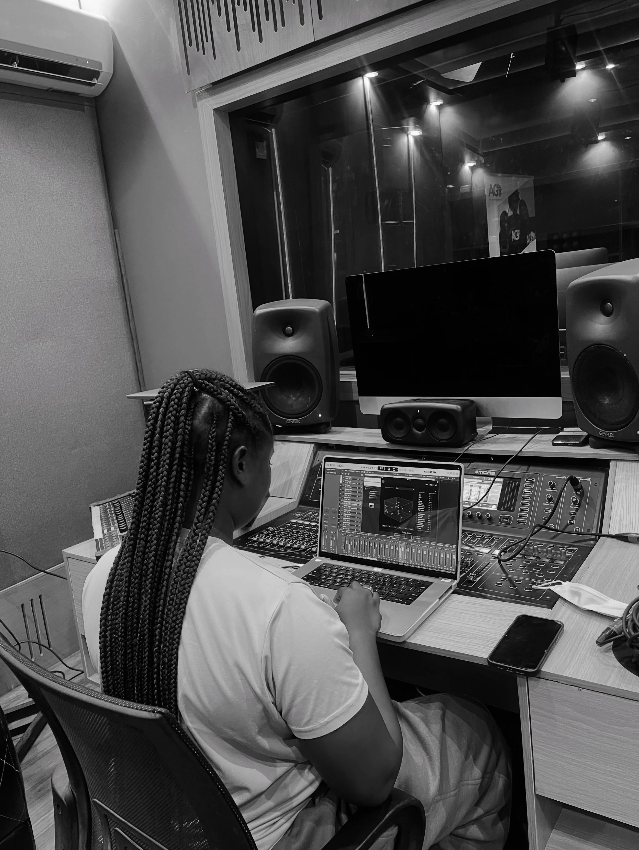 A person with braided hair working on a laptop in a music studio, surrounded by audio equipment and speakers.