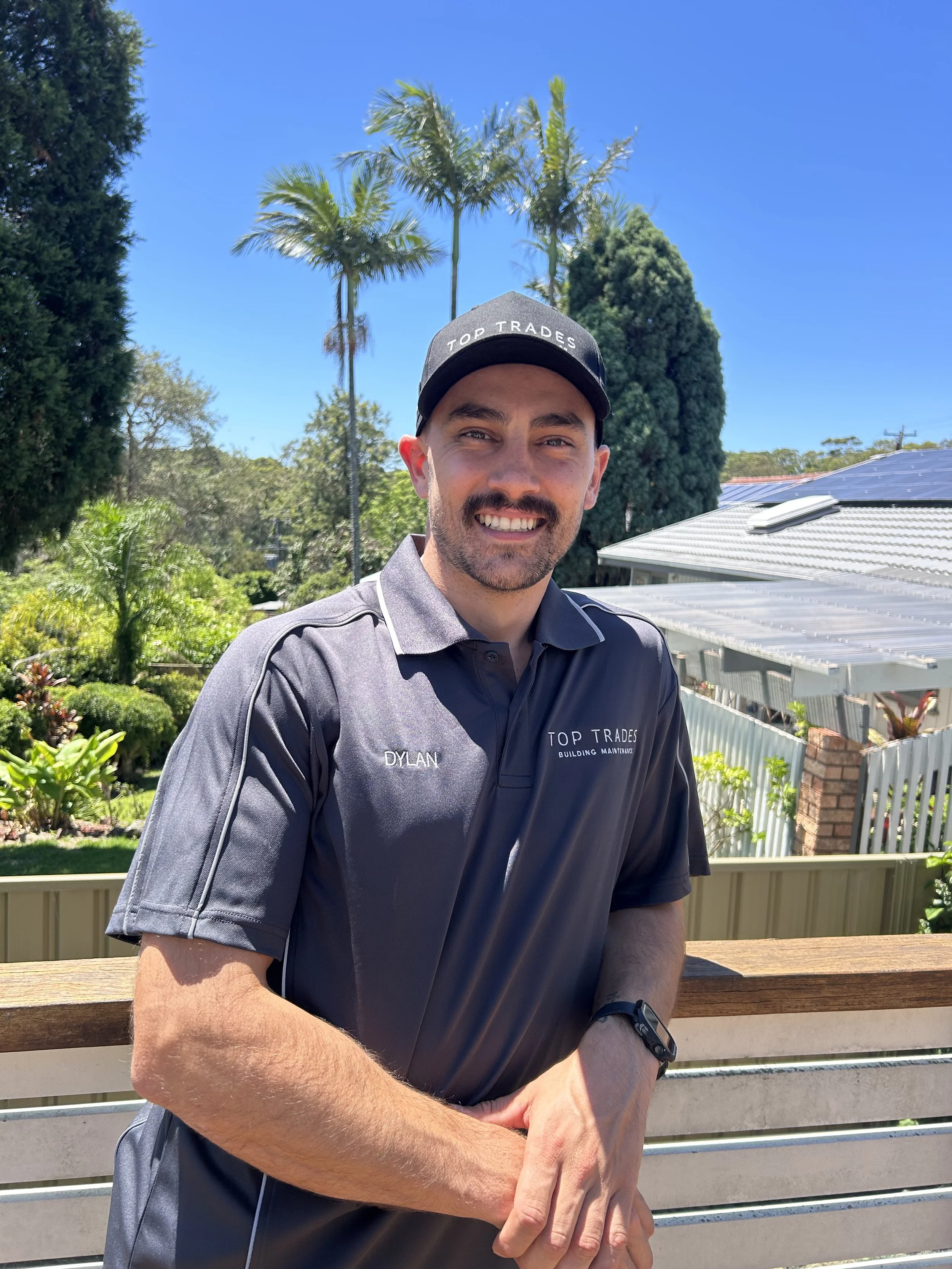 A smiling man outdoors on a sunny day, wearing a gray and black work shirt with 'Dylan' and 'Top Trades Building Maintenance' on it, a black cap with the same logo, a black watch, and standing in front of a wooden railing with lush greenery and tall palm trees in the background.