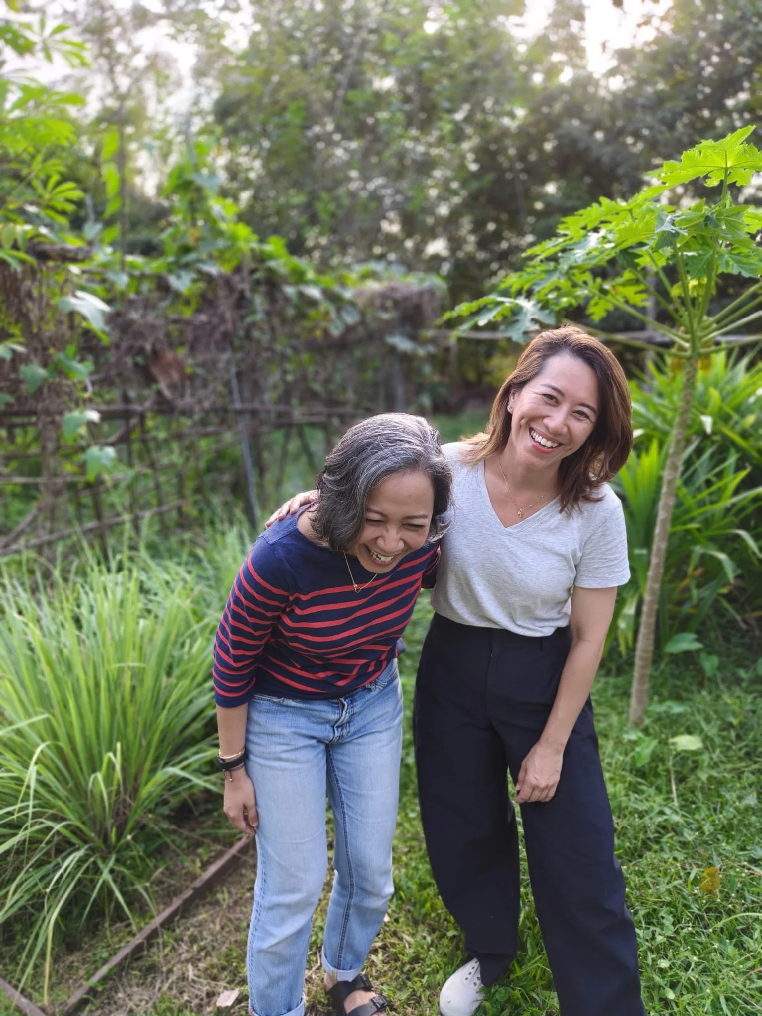 Two women smiling and laughing outdoors in a garden or park, surrounded by green plants and trees.