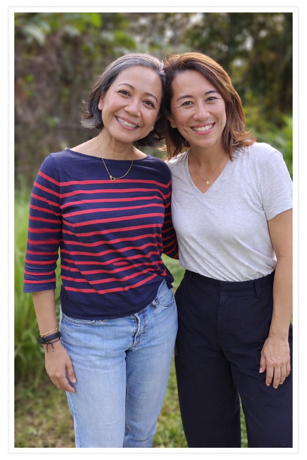 Two women smiling outdoors, standing close together, with trees and greenery in the background.