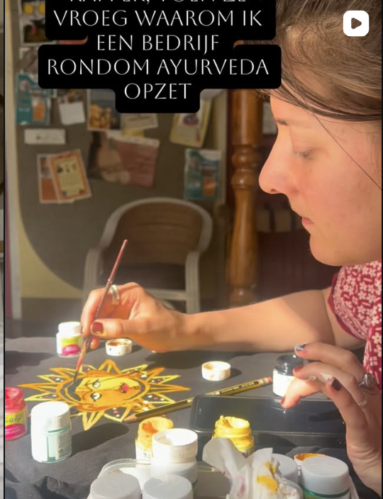 Een jonge vrouw schildert een zon met een gezicht op een tafel. Er zijn diverse potjes verf en een kwast op de tafel, en een achtergrond met een bord en een stoel.