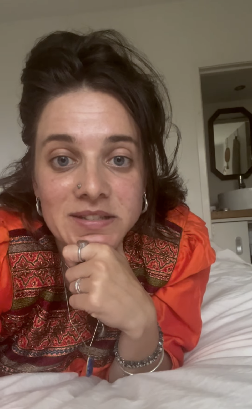 Vrouw met kort bruin haar, neus piercing, oorringen, sieraden, in een kamer met een wastafel op de achtergrond.