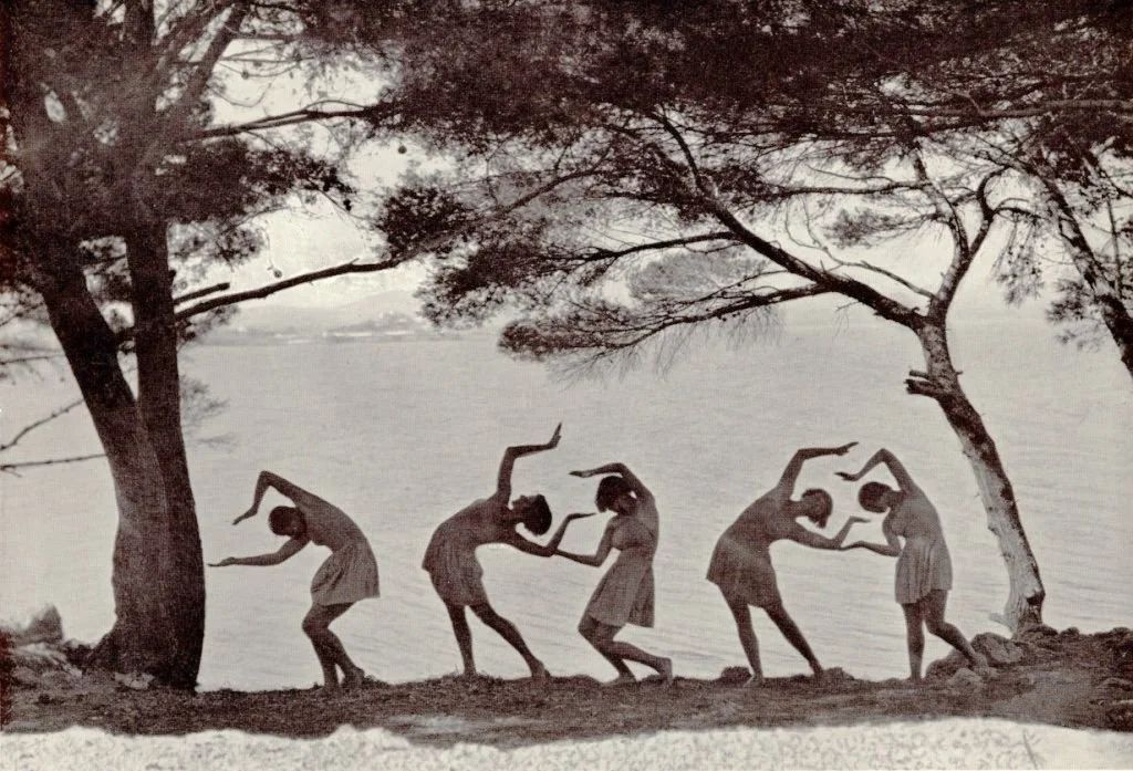 Zes vrouwen dansen in een rij onder bomen bij het water, de scène heeft een zwart-wit uitstraling.