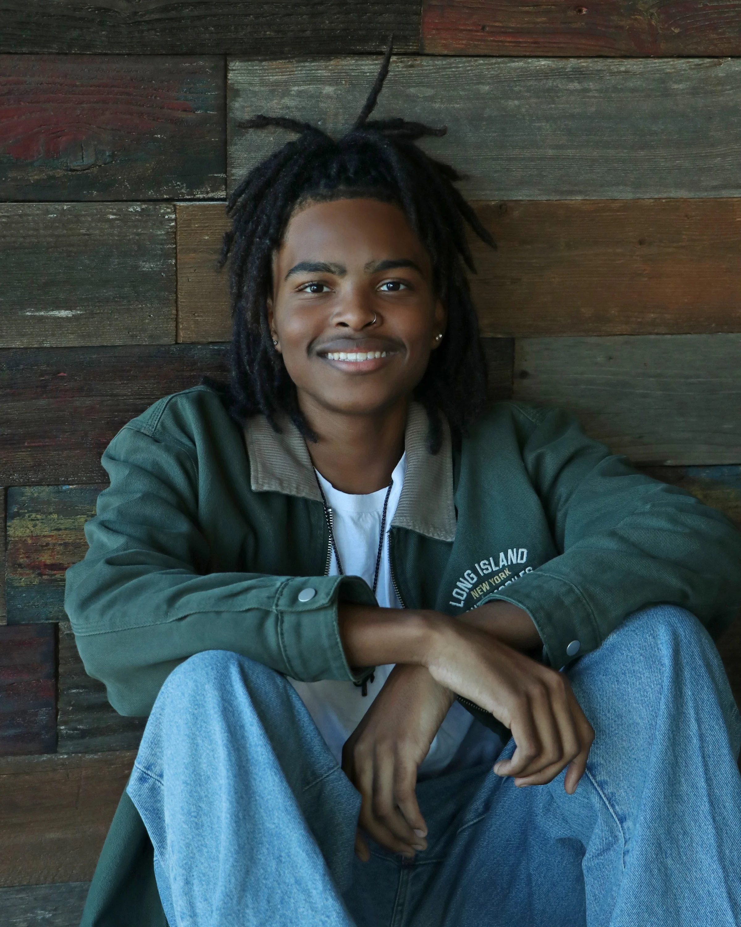 A person with medium skin tone and dreadlocks sitting against a wood-paneled wall, smiling, wearing a green jacket and blue jeans.