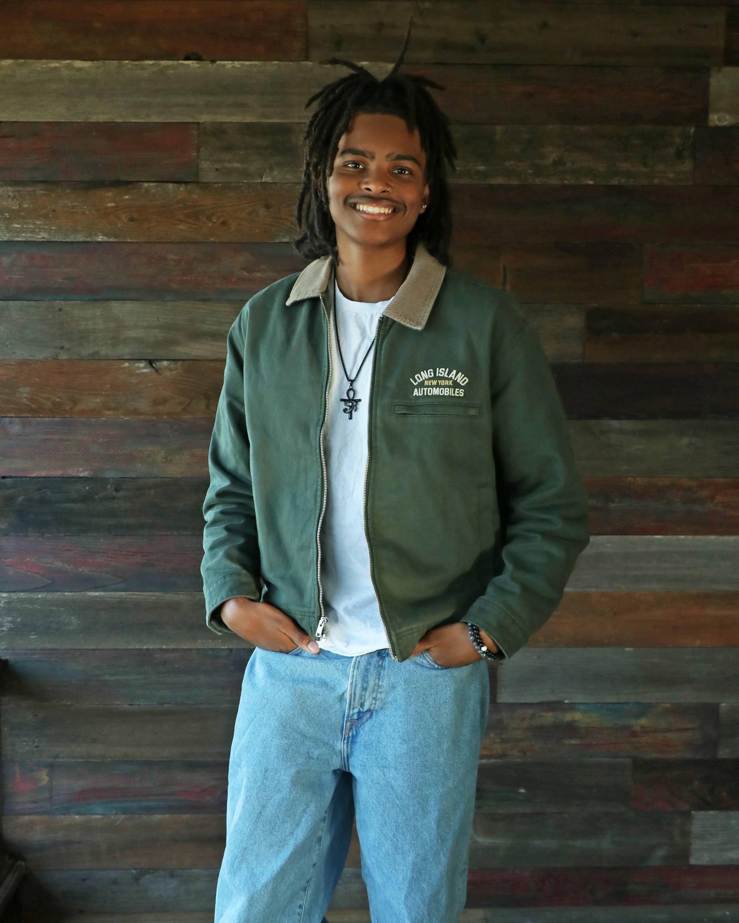Marcus Barrington, smiling, wearing a green jacket with 'Long Island Automobiles' logo, white shirt, jeans, and a cross necklace, standing in front of a wooden wall.