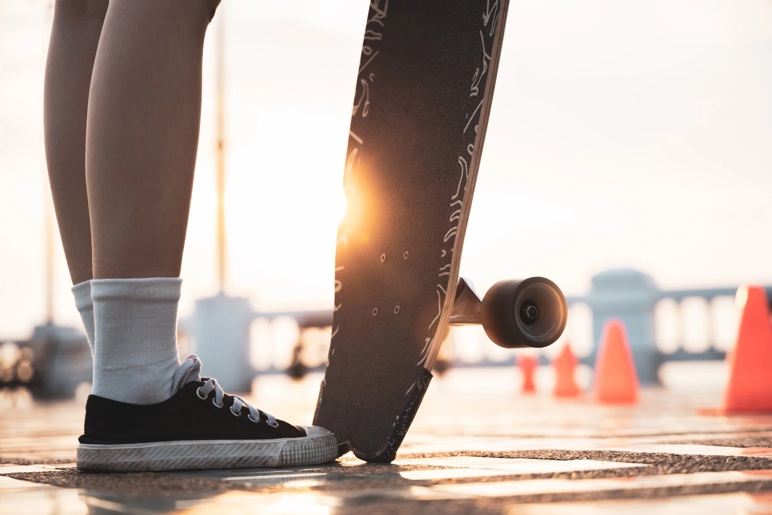 Person wearing black sneakers and white socks standing next to a skateboard on a wooden surface during sunset with traffic cones in the background.