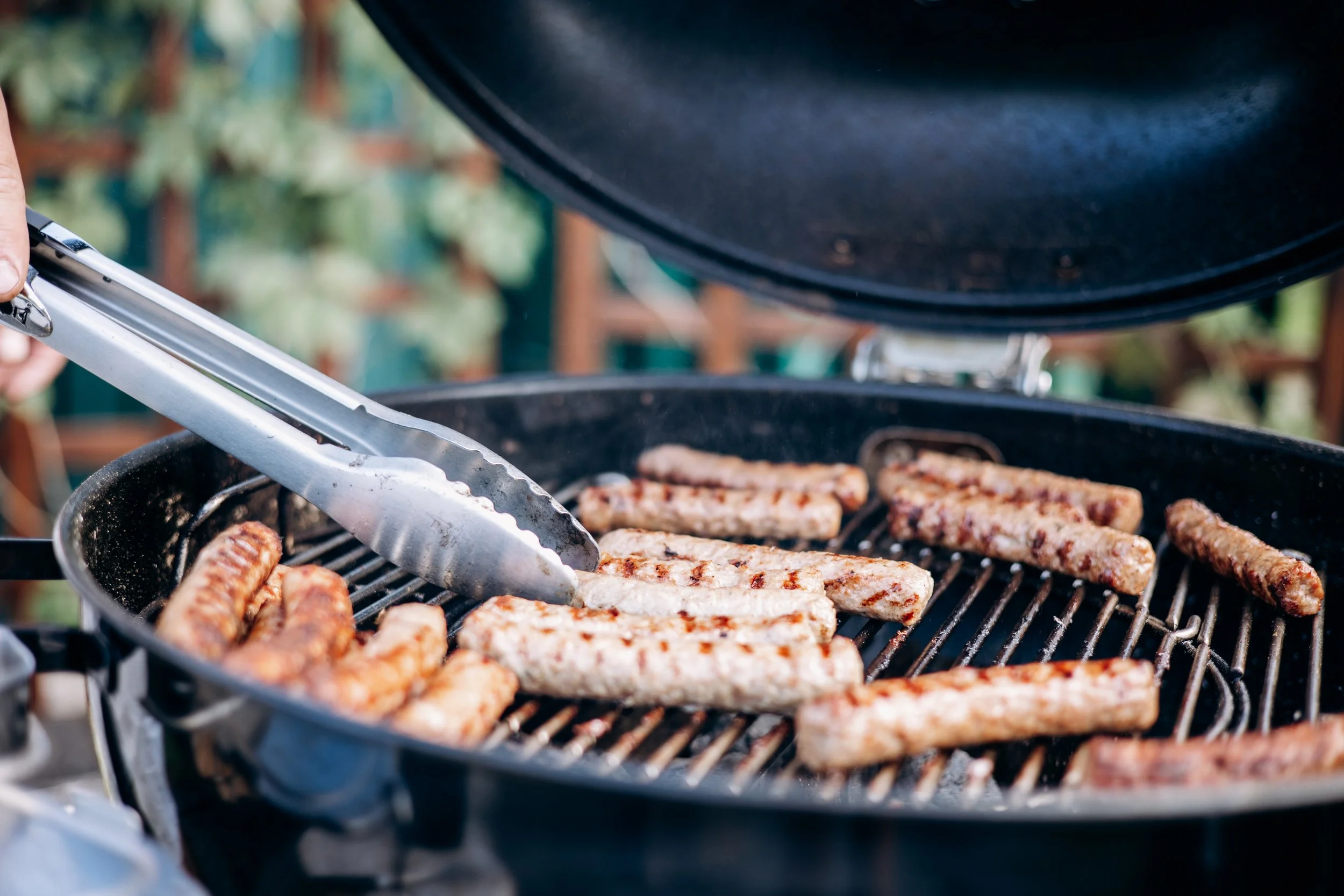 Meat being grilled on an outdoor barbecue, with tongs gripping some meat strips on the grill.