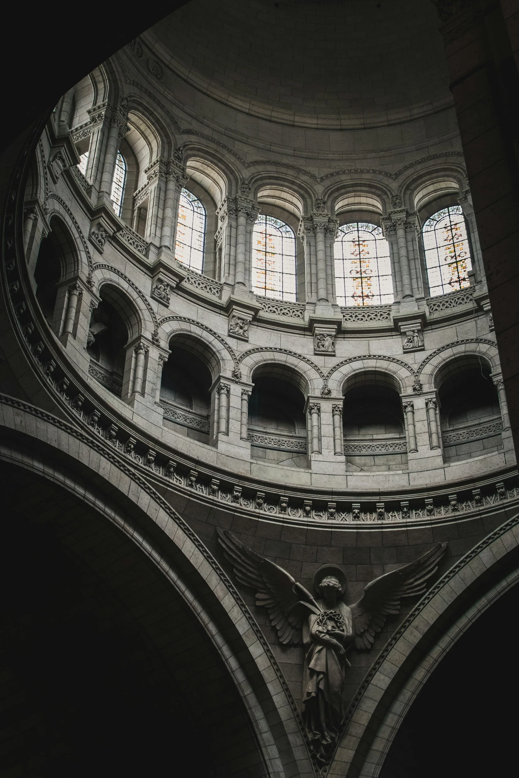 Interior of a domed building with intricate stone architecture, stained glass windows, and a sculpture of an angel with wings holding a shield.