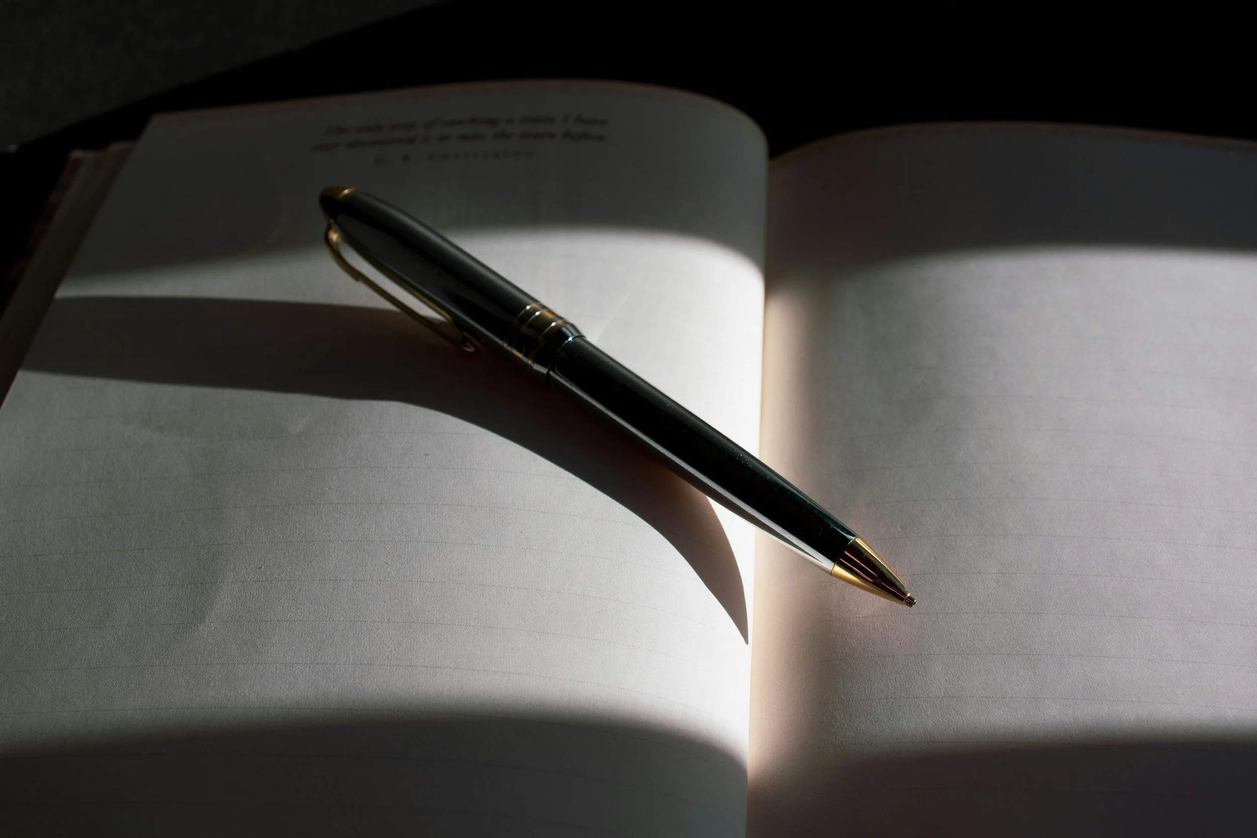 Open notebook with lined pages and a black and gold pen resting on it, casting a shadow in a dimly lit environment.