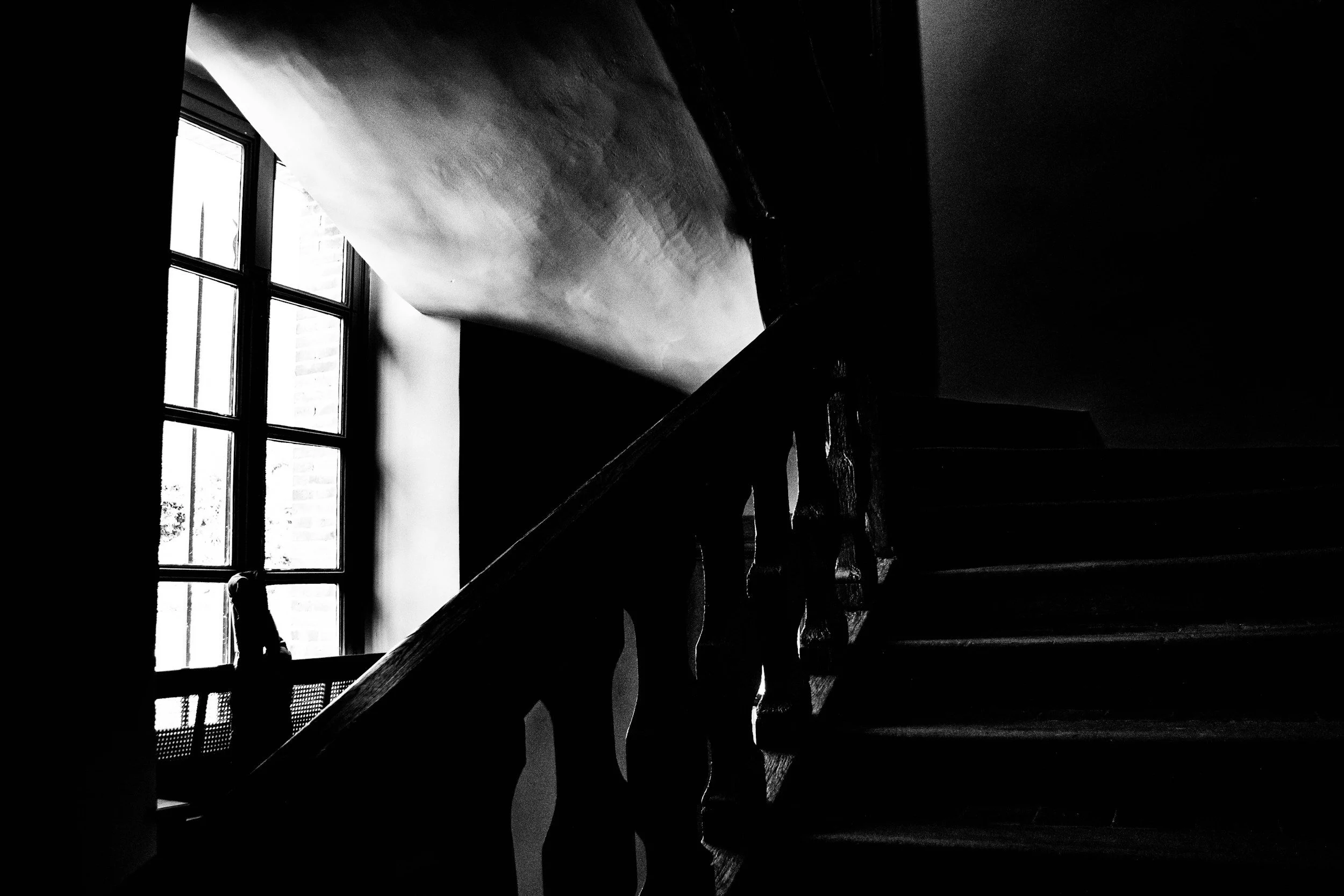 Black and white photo of a staircase next to a large window with multiple panes, showing a silhouette of a person sitting at a table near the window.