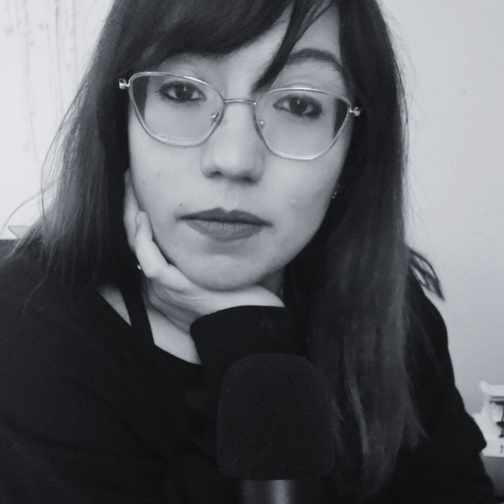A woman with shoulder-length dark hair and glasses posing with her hand resting on her chin in a black and white photo.