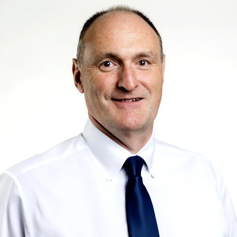 A middle-aged man with short brown hair, wearing a white dress shirt and a dark blue tie, smiling at the camera against a plain white background.