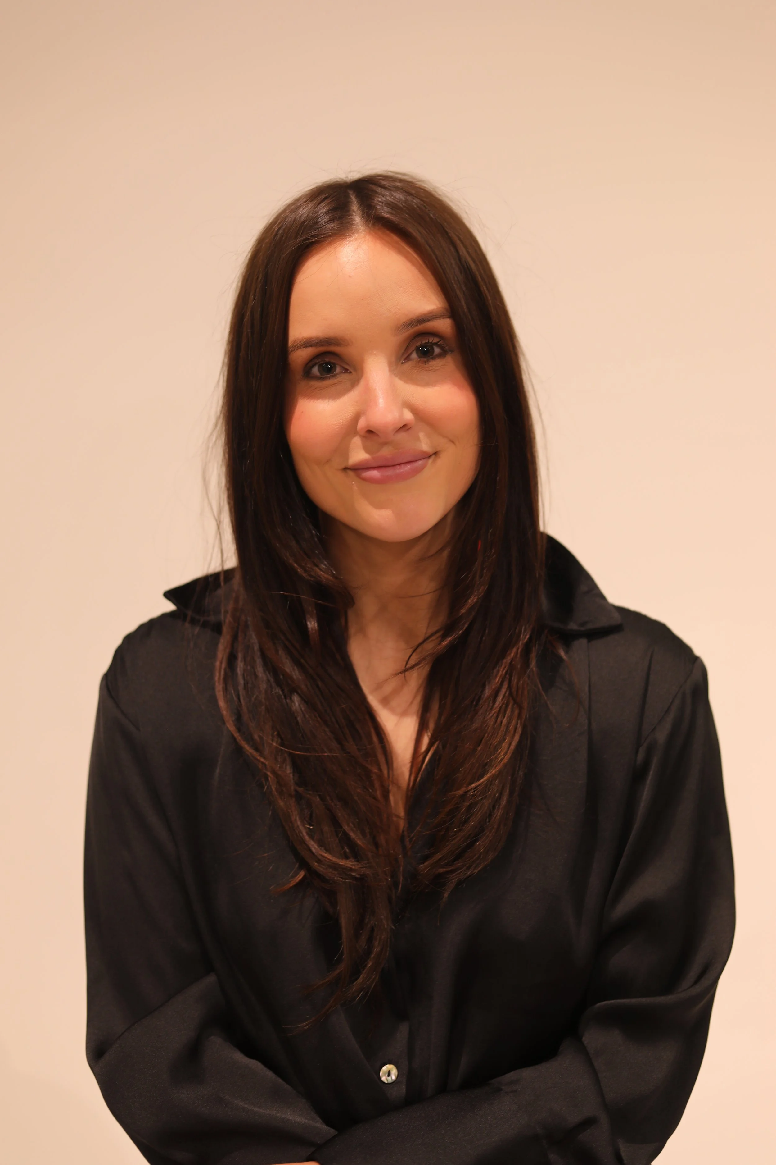 A woman with long brown hair smiling, wearing a black blouse, standing against a plain light-colored background.